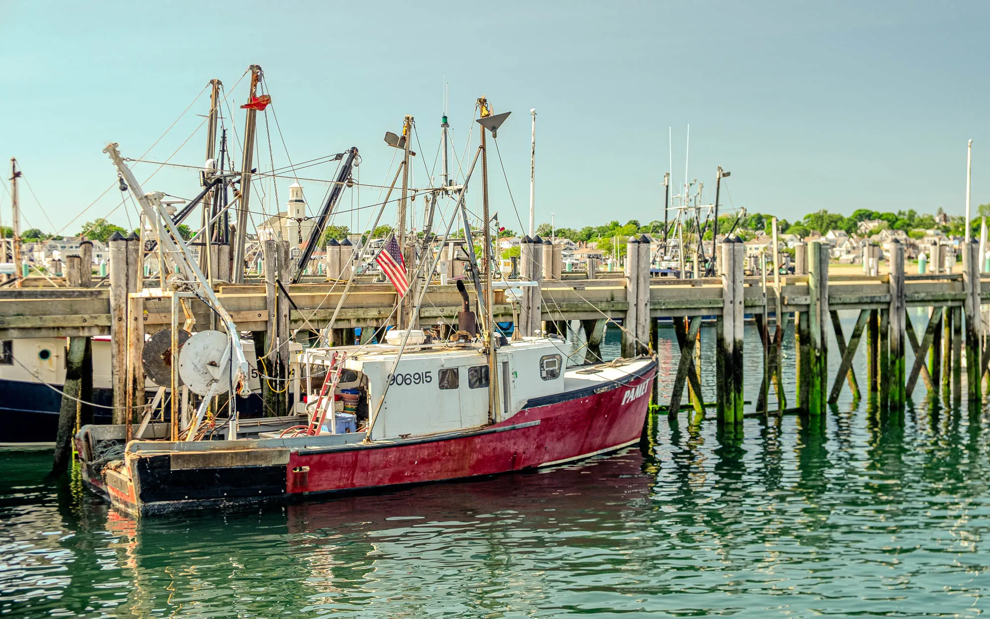 A weathered fishing boat, primarily red and white, is docked at a wooden pier on a sunny day. The boat, named "PAMELA", is positioned with its bow pointing towards the right, partially obscuring the dock behind it. The hull is a deep, faded red, showing signs of wear and age, while the cabin is mostly white with visible numbers and a small window. A small American flag is attached to one of the boat's many masts and rigging. These masts and rigging, made of metal and rope, reach upwards towards the clear, bright sky, creating a complex network of lines. The wooden pier is constructed with weathered planks and support beams, some of which are partially submerged in the green-tinged water. In the background, a small town or village can be seen along the shoreline, featuring light-colored buildings and trees, partially obscured by the masts and rigging of other boats. The water reflects the colors of the sky and surroundings, creating shimmering patterns.