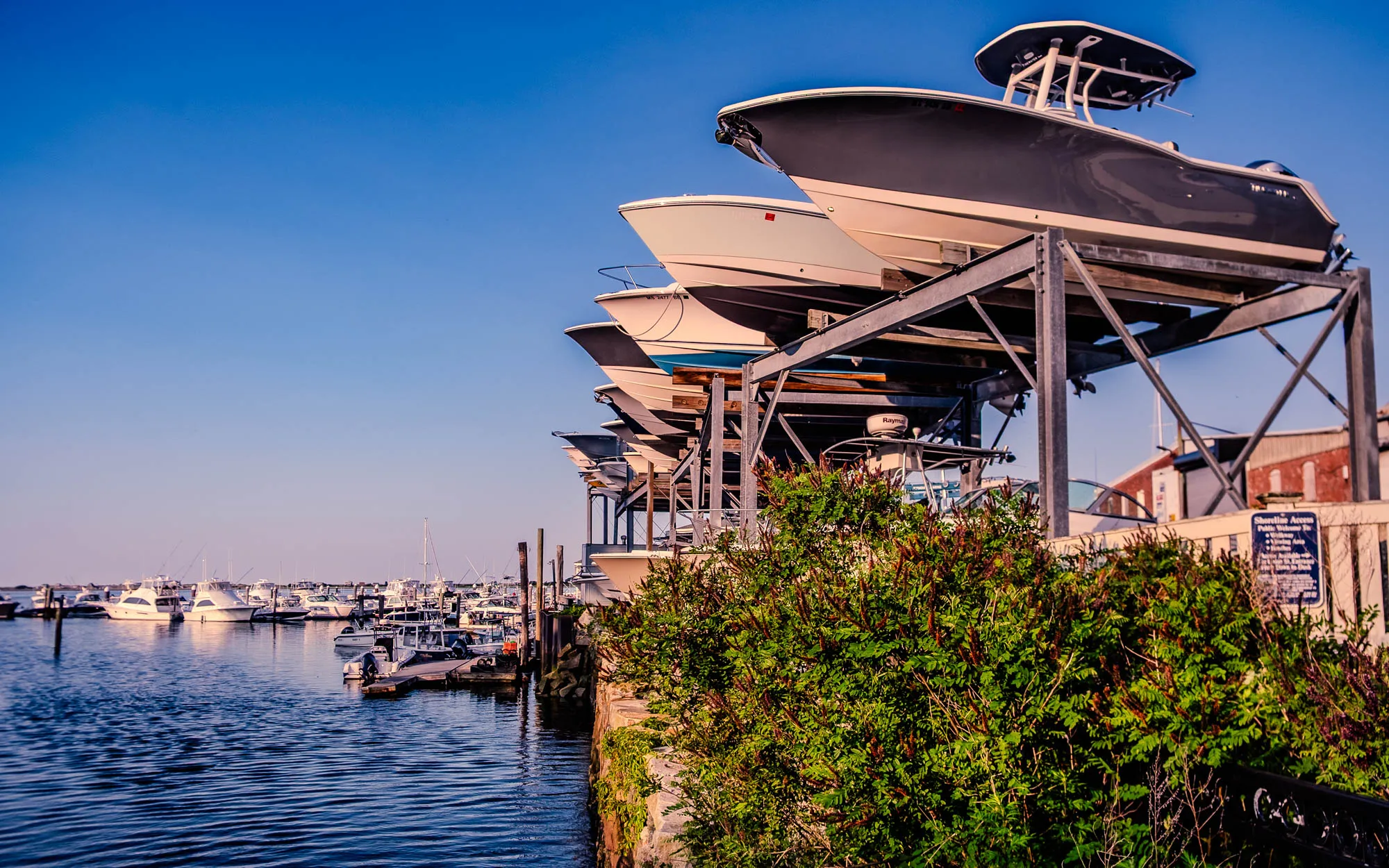 The image shows a marina with several boats docked and a boat storage rack filled with boats. In the foreground, dark blue water ripples gently. Along the shore, a low wall supports vibrant green bushes with reddish-brown spikes. Behind the bushes, a gray metal rack holds several boats stacked on top of each other. The boats on the rack are various sizes and colors, ranging from white to gray, and are arranged in a descending order, with the largest boat at the top. In the background, more boats are docked along a pier, creating a busy marina scene. A clear blue sky fills the upper portion of the image, suggesting a bright and sunny day.