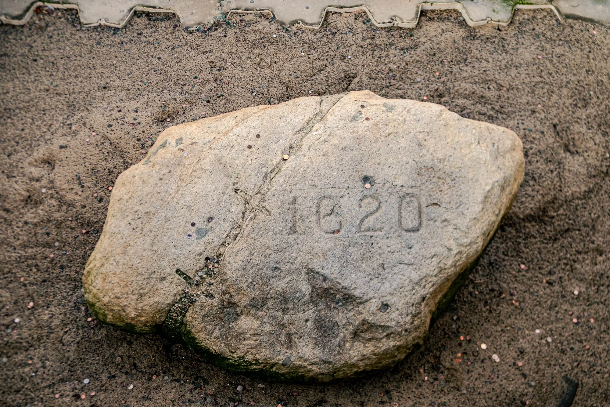 The image shows a tan-colored, roughly rectangular stone resting on a surface of brown, sandy soil. The stone appears weathered, with variations in texture and color across its surface. Carved into the stone, in slightly recessed numerals, is the year "1620." The carving is somewhat faded, blending in with the stone's overall color. Several cracks or fissures run across the stone's surface. Scattered around the stone, both on its surface and in the surrounding soil, are numerous small, shiny coins. These coins are predominantly copper-colored, but some may be silver or gold-toned. The soil around the stone is grainy and appears moist. At the top of the image there is a concrete edge that has several coins on it, as well as spots of moss.