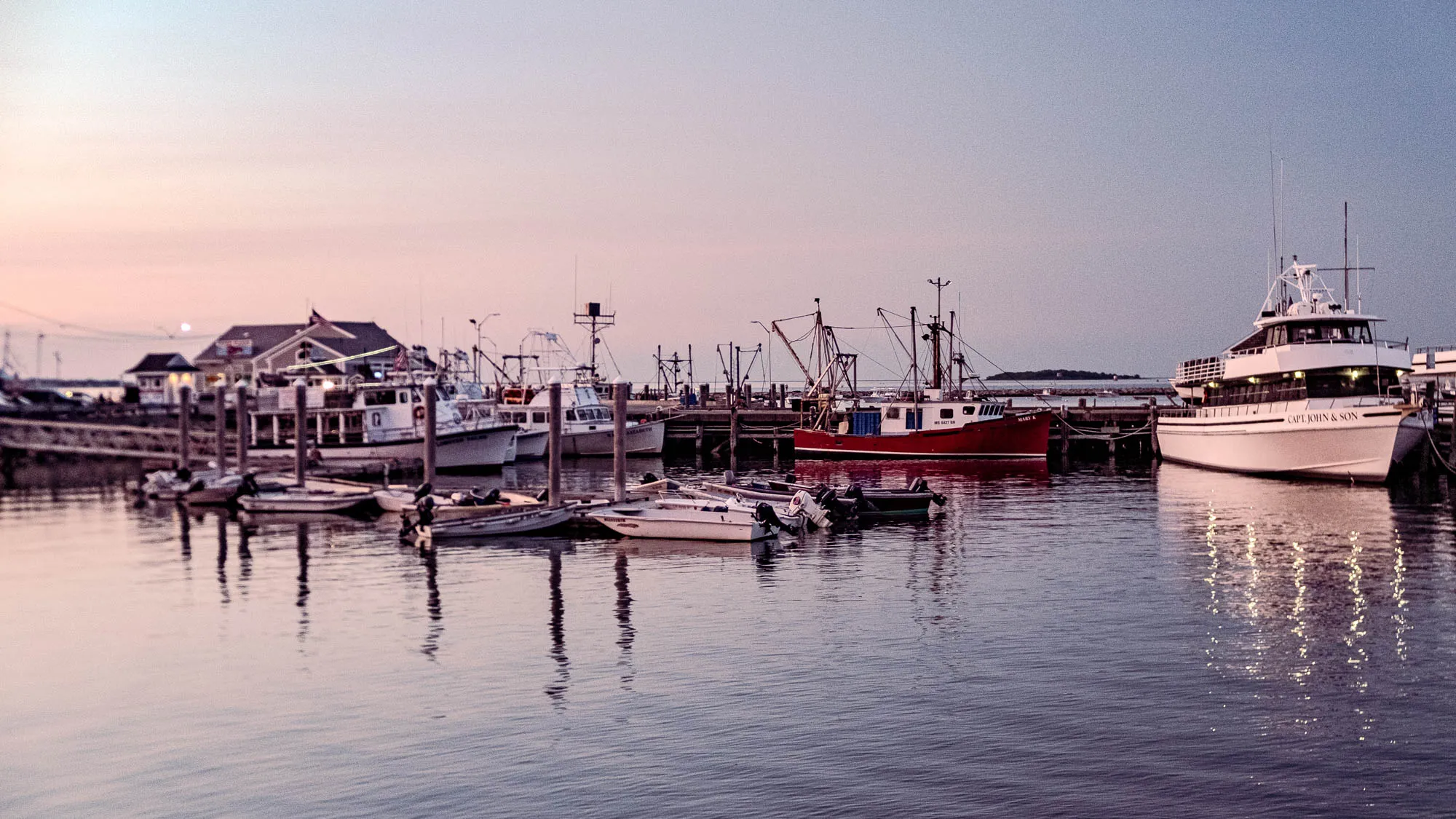 The image shows a tranquil harbor scene at twilight. Several boats of varying sizes are docked alongside a pier. The sky transitions from a soft, pale pink near the horizon to a muted blue higher up. The water is calm, reflecting the colors of the sky and the lights from the boats and buildings, creating a shimmering effect. In the foreground, small motorboats are clustered together, gently bobbing in the water. A red fishing boat stands out among the others, with its rigging silhouetted against the sky. Further to the right is a larger, white yacht with the words 'CAPT. JOHN & SON' printed on its side. Along the pier, a couple of buildings are visible, including one with an American flag waving from its roof. The overall scene evokes a sense of peace and quiet.