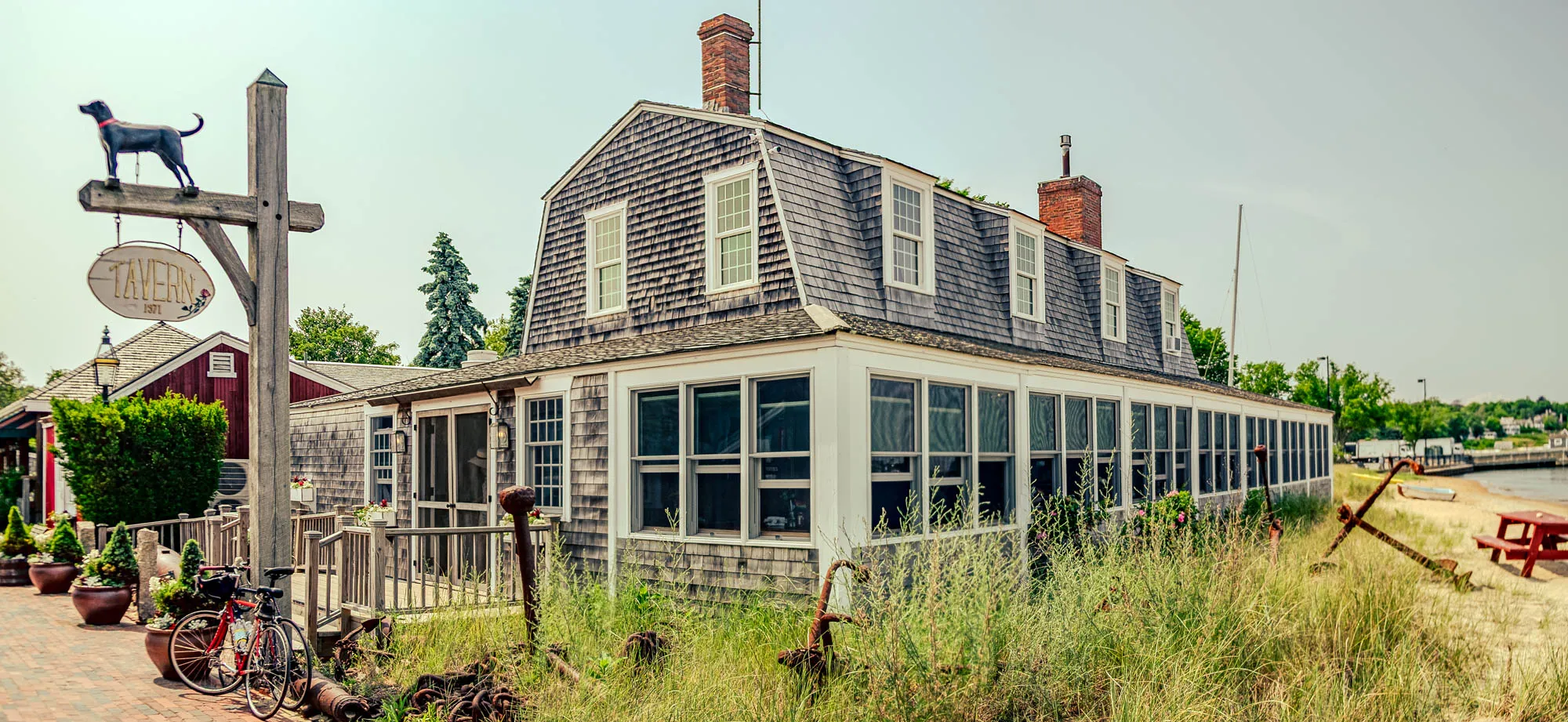 The image shows a charming, weathered tavern nestled in a coastal landscape. The main building is covered in gray shingles and features a steeply pitched roof with multiple dormer windows, each framed in white. A long, glass-enclosed porch extends along one side, allowing plenty of natural light inside. The porch windows offer a glimpse of the interior, but details are obscured. In the foreground, tall beach grass and various nautical artifacts, including rusty anchors and chains, add to the seaside atmosphere. To the left, a weathered wooden signpost displays a dark silhouette of a dog atop the words 'Tavern 1871', painted in a vintage style. Behind the tavern, a glimpse of a small beach with a red picnic table and boats resting on the sand can be seen, hinting at the tavern's proximity to the water. The sky is a hazy, bright blue, suggesting a warm, sunny day.