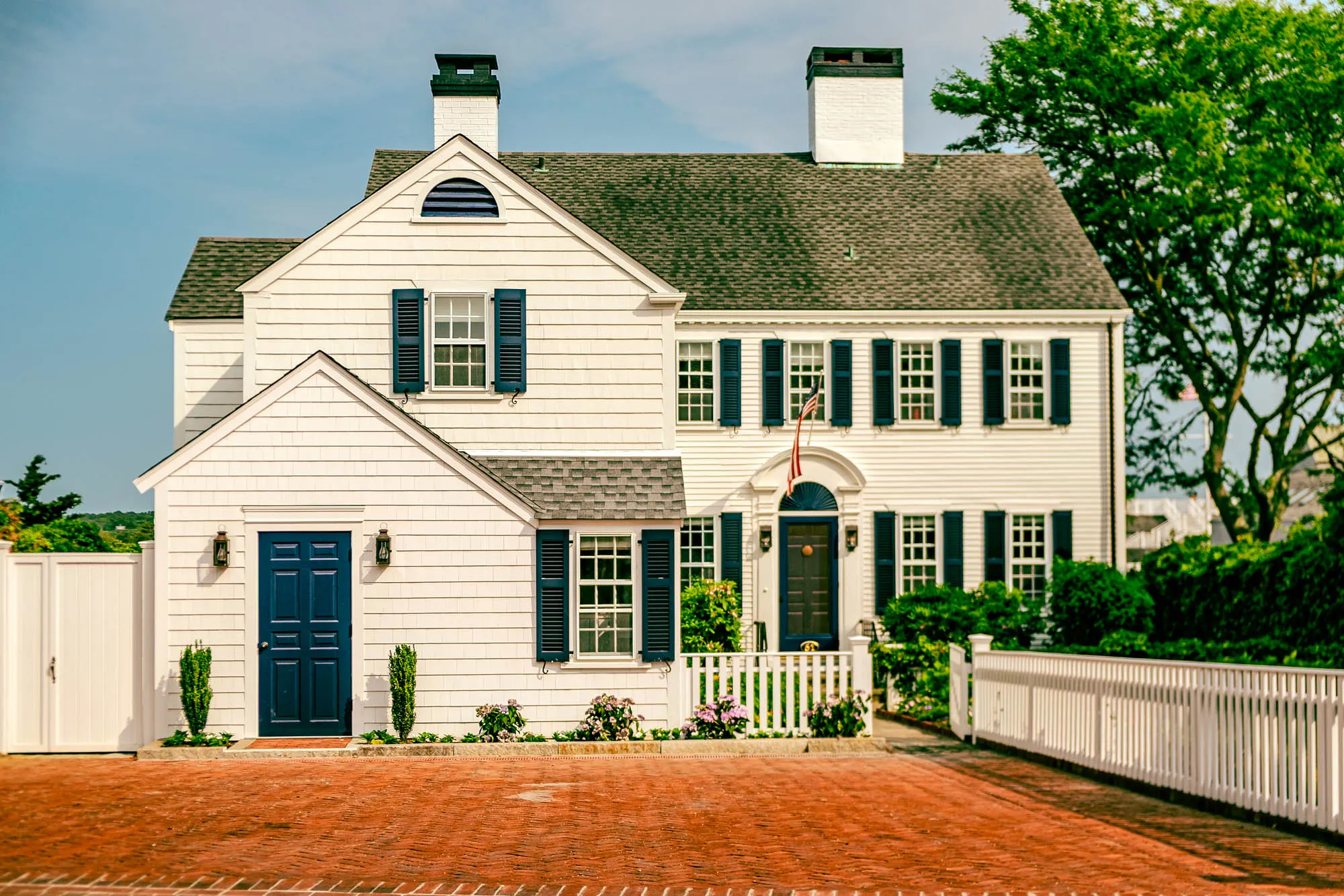 A stately two-story house with white clapboard siding stands prominently against a pale blue sky. The house features a dark gray shingled roof and two white chimneys with black tops. Dark blue shutters adorn the numerous windows, adding contrast to the bright facade. A smaller, two-story section is attached to the main structure on the left, with a striking dark blue door flanked by sconces and trimmed hedges. A quaint white picket fence encloses the front of the property, partially obscured by lush greenery and flowering plants. A brick walkway leads to the house, creating a warm, inviting entrance. An American flag hangs over the main entrance. A large, mature tree with vibrant green foliage partially frames the right side of the house, adding a touch of nature to the scene.