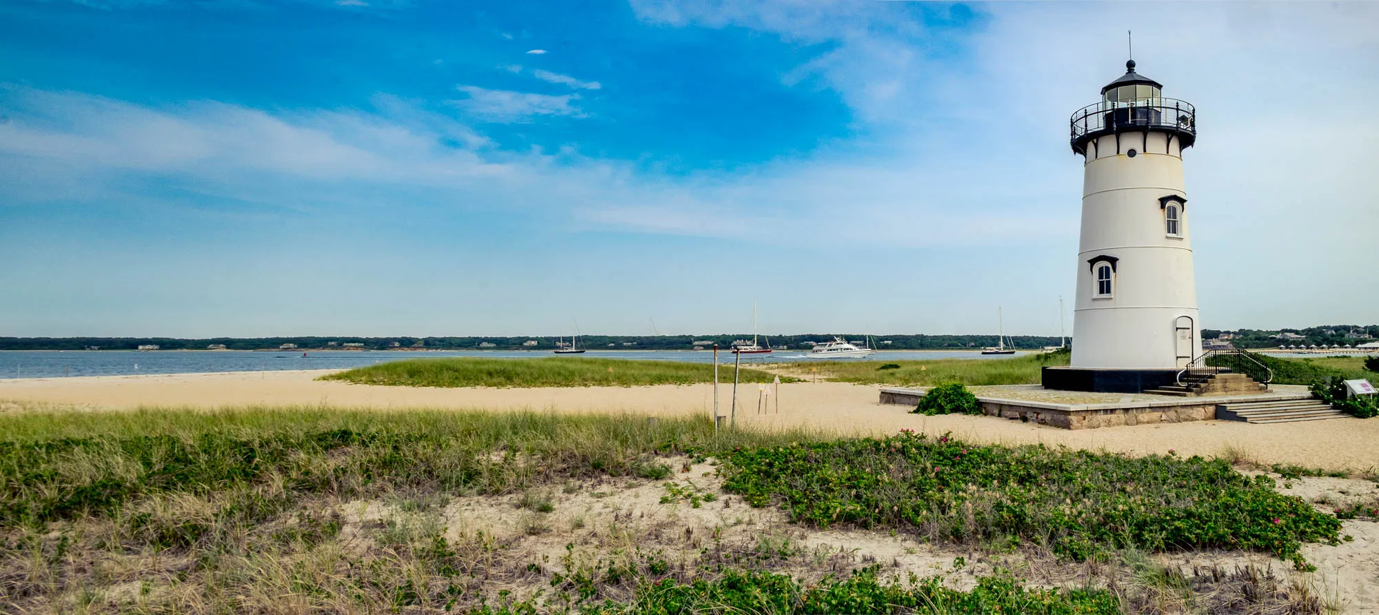 The photograph features a scenic coastal landscape dominated by a tall, white lighthouse. The lighthouse stands prominently on the right side of the image, featuring a black lantern room at its top with a railing. Its white structure has small, dark rectangular windows and a black door. To the left of the lighthouse, a sandy beach stretches towards a calm body of water. Low sand dunes covered in beach grass run along the foreground and middle ground. In the distance, across the water, a shoreline is visible, marked by trees and buildings. Several sailboats and a larger motorboat are scattered on the water, adding to the maritime feel. The sky above is a bright blue, dotted with soft, wispy clouds, suggesting a sunny, clear day.
