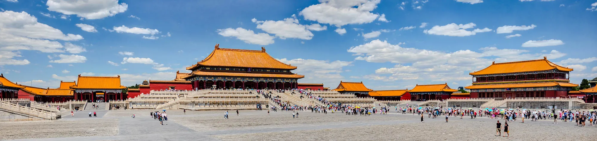 The image shows a wide panoramic view of a large courtyard in front of the Forbidden City in Beijing, China.  The sky is bright blue with fluffy white clouds, and the ground is a light gray stone.  There are many people walking around the courtyard, and a large red and gold building is in the middle of the image.  The building has many roofs, and the eaves are decorated with intricate carvings.  There are other buildings in the background, some of which are also decorated with red and gold.  The image is bright and cheerful, and it gives a sense of the grandeur of the Forbidden City. 
