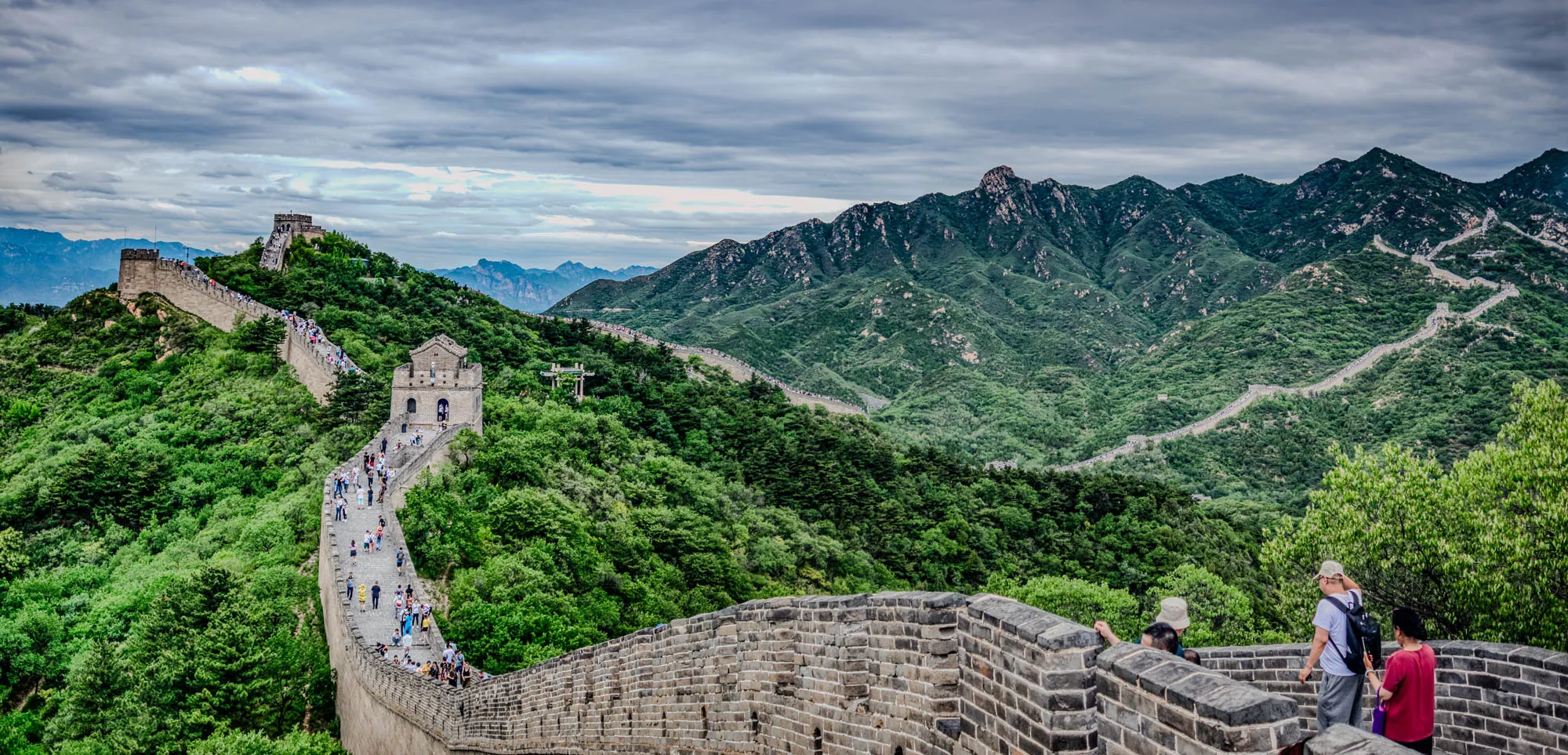 The image shows a view of the Great Wall of China. The wall is a long, winding structure made of stone. It stretches across a mountain range and is covered in green trees and bushes. In the foreground, there is a group of people standing on the wall, looking out at the scenery. There is a cloudy sky in the background. The Great Wall is a UNESCO World Heritage Site and is one of the most popular tourist destinations in China. The wall is a symbol of the country's history and culture.