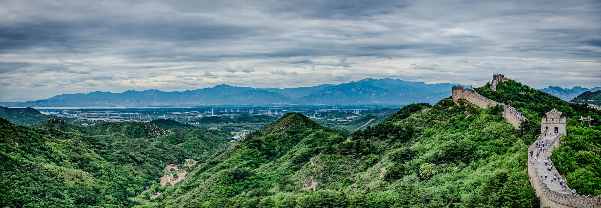 The Great Wall of China winds its way through the landscape of rolling hills and valleys. The wall is made of stone and has a pathway along the top that people are walking on. The wall is surrounded by lush green trees and foliage. In the distance, a row of mountains can be seen against a cloudy sky. The image is taken from a high vantage point, looking down onto the wall and the surrounding landscape. The sky is overcast with a few patches of blue visible through the clouds.