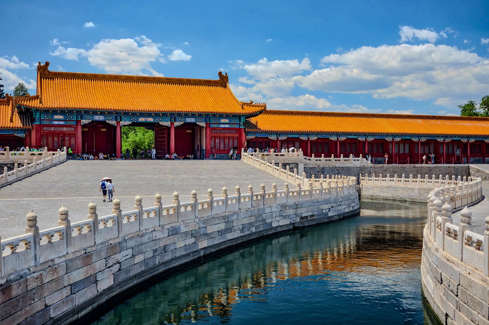 The image shows a view of a large, traditional Chinese palace complex. The photo was taken from a slightly elevated point of view, looking down onto the courtyard. In the foreground, there is a stone bridge spanning a moat filled with still, blue water.  The bridge is lined with a low white stone railing. To the left of the bridge, two people are walking away from the viewer, the person in front is holding an umbrella. To the right of the bridge, there is another stone wall, curving away from the viewer. On the other side of the moat, there is a large complex of buildings with red pillars and golden roofs. The buildings appear to be at least two stories tall and have many windows and doorways. The roof is adorned with intricate details in blue and green. The sky above the complex is a bright blue with white clouds.