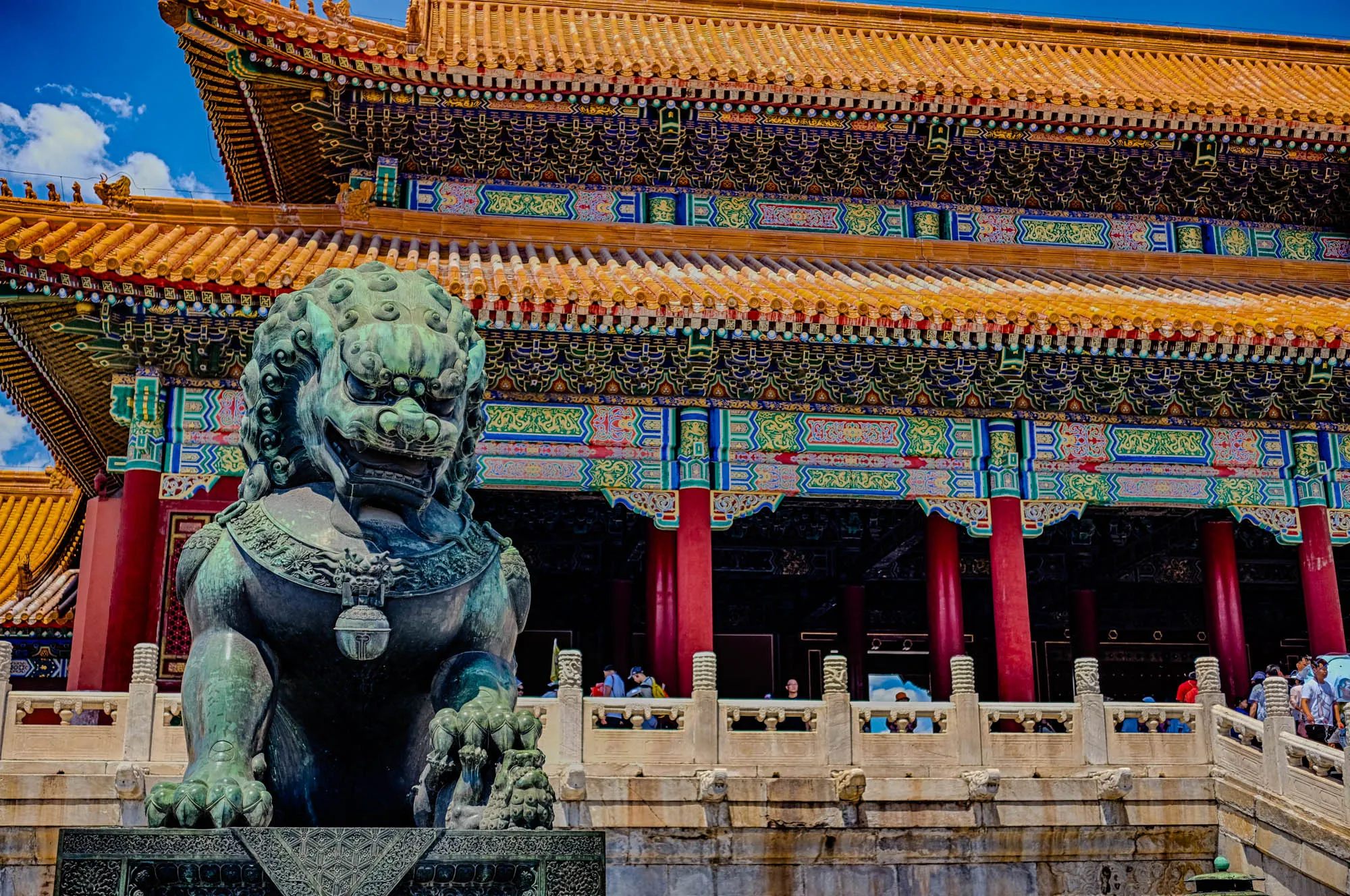 The image shows a large, green, stone Chinese lion statue in front of a brightly colored, ancient Chinese temple. The lion is sitting on its haunches with its front paws outstretched. It appears to be guarding the entrance to the temple. The temple is adorned with elaborate details in red, gold, blue, and green. The roof is tiled with a repeating pattern of yellow tiles and small, red, decorative elements. The temple is surrounded by a stone wall and a large, open courtyard. The image is likely taken from a low angle, looking up towards the temple.