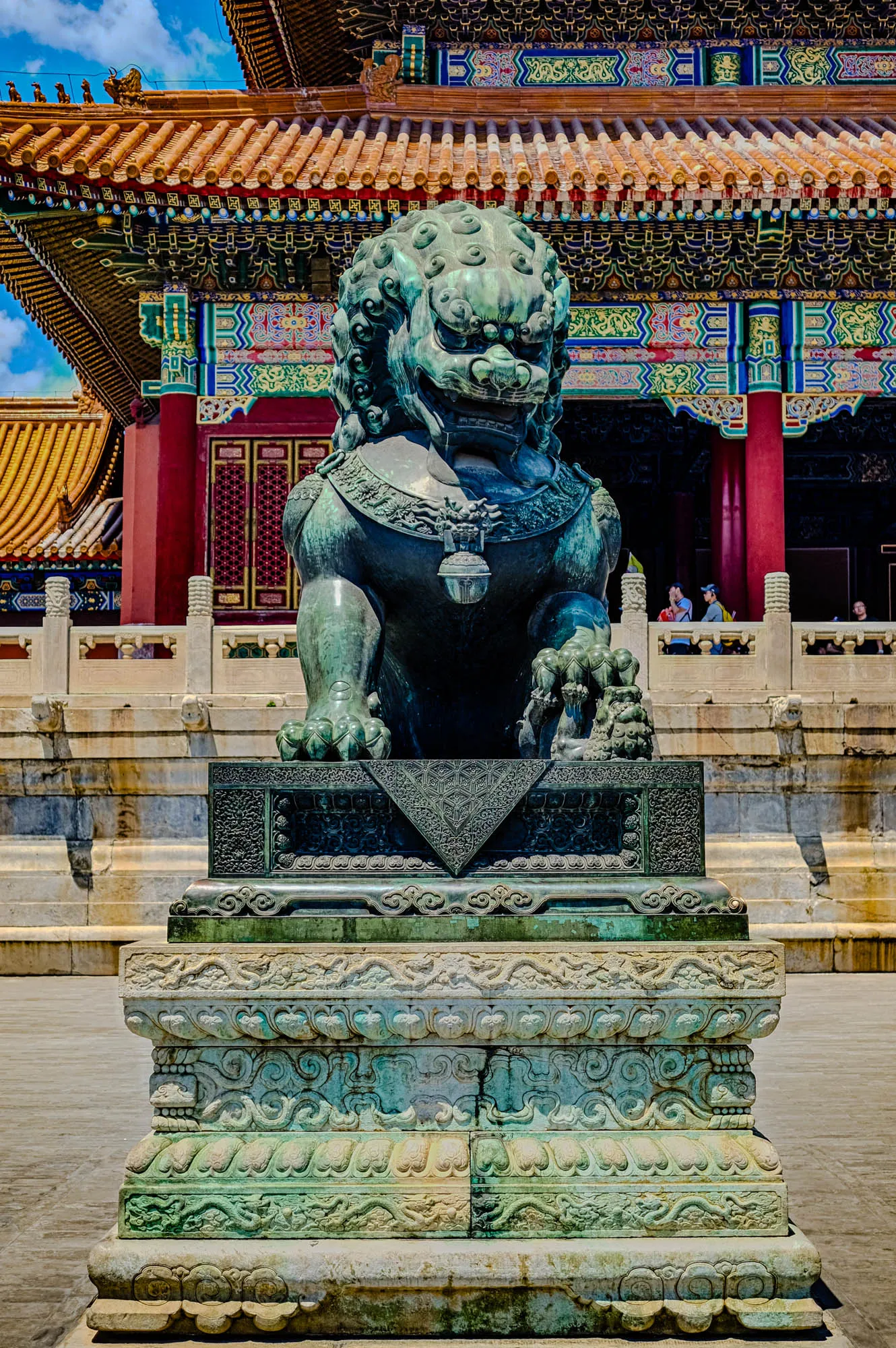 The image shows a green stone Chinese lion statue standing on a multi-tiered, intricately carved stone base. The lion is facing forward with its mouth slightly open, and its body is covered in detailed carvings. The lion is standing on a platform that is surrounded by a stone railing. In the background, there is a large building with a red roof and a colorful, ornate facade. The building appears to be a traditional Chinese structure. There are a few people walking around the building and platform, and the sky is a clear blue with a few clouds.