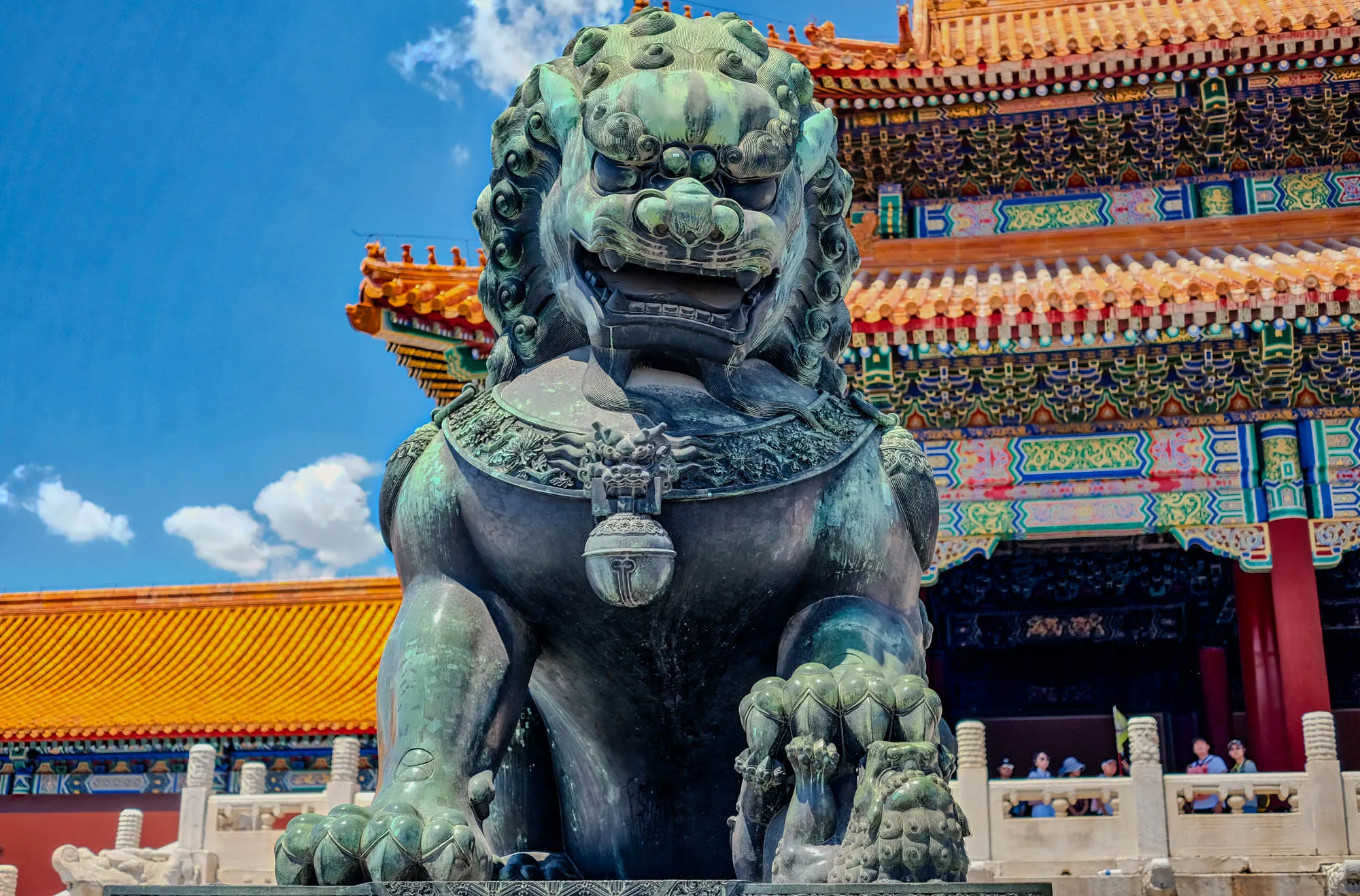 This is a photo of a large, jade lion statue. The lion has a fierce expression, with a wide mouth and large, sharp teeth. Its left paw is standing on a cub, looking up. Its body is covered in intricate carvings. It is sitting with its front legs extended, and its back legs are tucked underneath its body. 

The lion is standing in front of a colorful, ornate building with a red roof. The building is decorated with elaborate carvings and paintings. There are people standing on a balcony in front of the building. The sky is blue and cloudy, and there is a hint of a bright orange roof in the background. 
