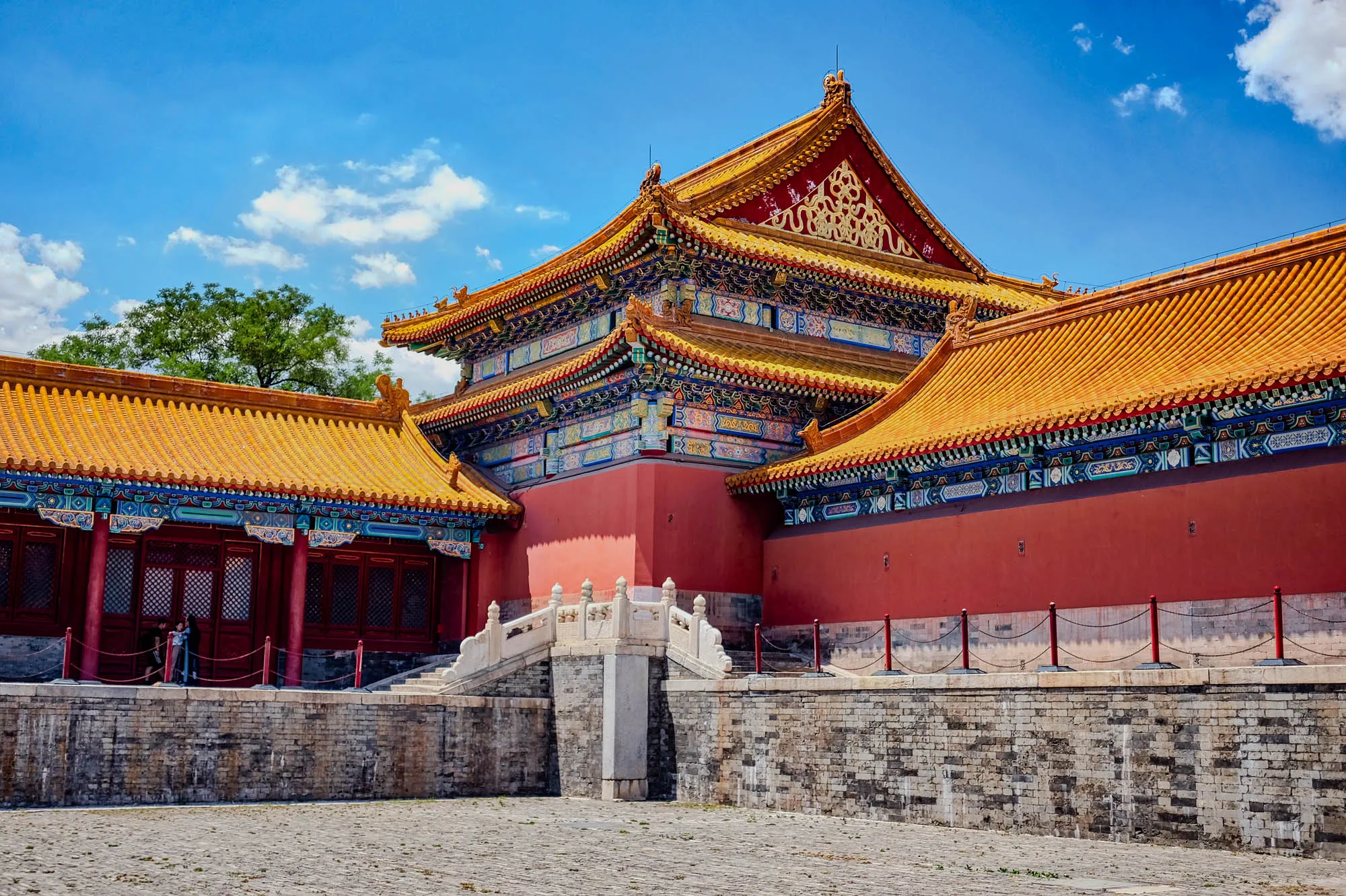 The image shows a large, ornate building with multiple tiers and a red roof. The roof is covered in golden tiles and has a series of intricate carvings and designs, including swirling patterns and geometric shapes. The building has a series of pillars and arches, and there is a stone staircase leading up to the entrance. The building is surrounded by a stone wall and there are several people standing on the courtyard in front of the building. The sky is blue and there are some clouds in the distance. The building is likely a traditional Chinese temple or palace.