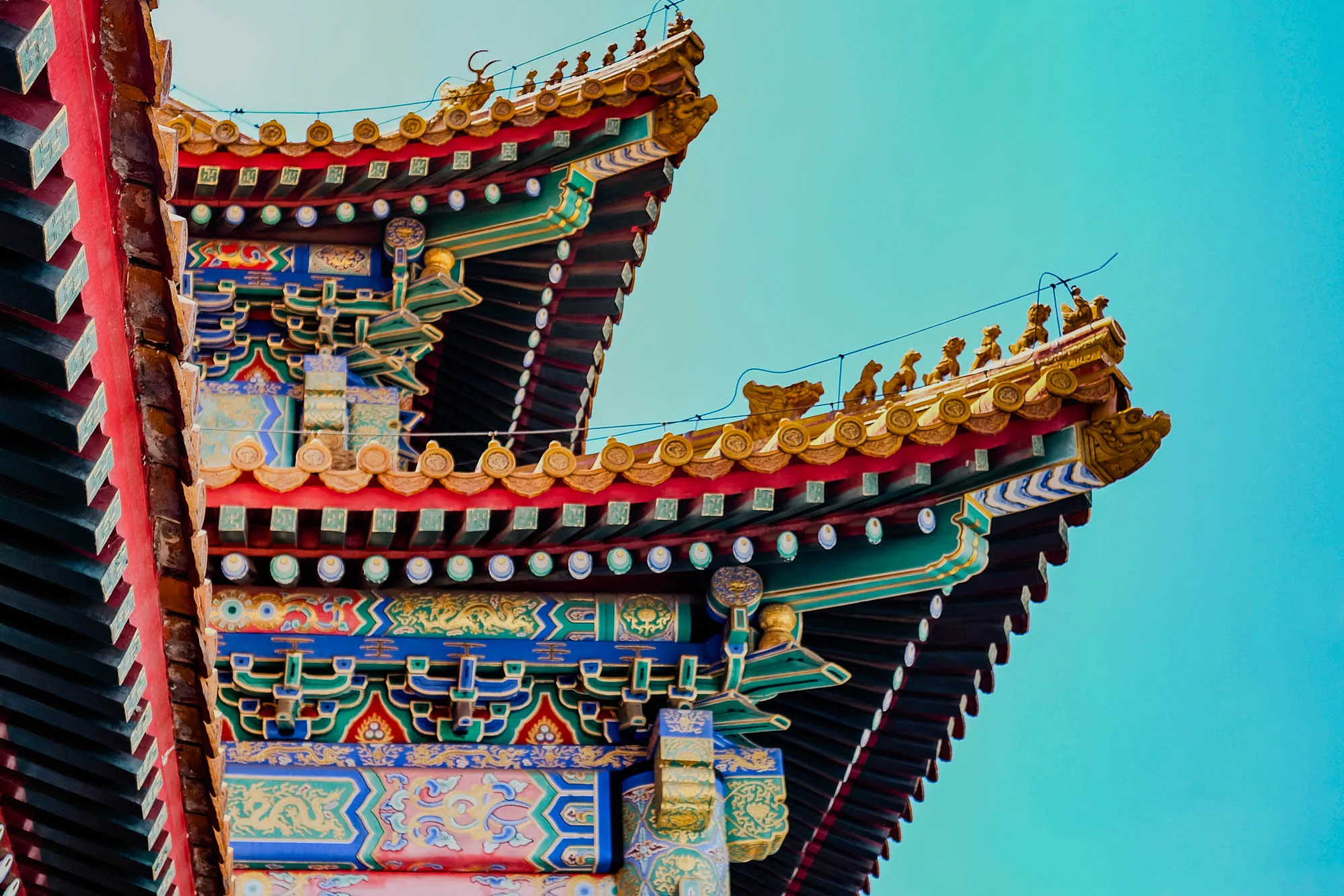 The image shows a close-up of a brightly colored, intricately carved, and decorated Chinese temple roof against a bright blue sky. The roof is layered, with multiple tiers of wooden beams painted in black, red, green, and gold. The top tier is decorated with gold mythical beasts and a row of gold circles. Underneath are more gold circles, alternating with green circles.  The next tier features a row of green and black squares, followed by a layer of vibrant blue, green, and gold floral designs. The roof is accented with red trim and has a slightly curved, upward slant. The overall effect is one of impressive craftsmanship and traditional Chinese artistry.