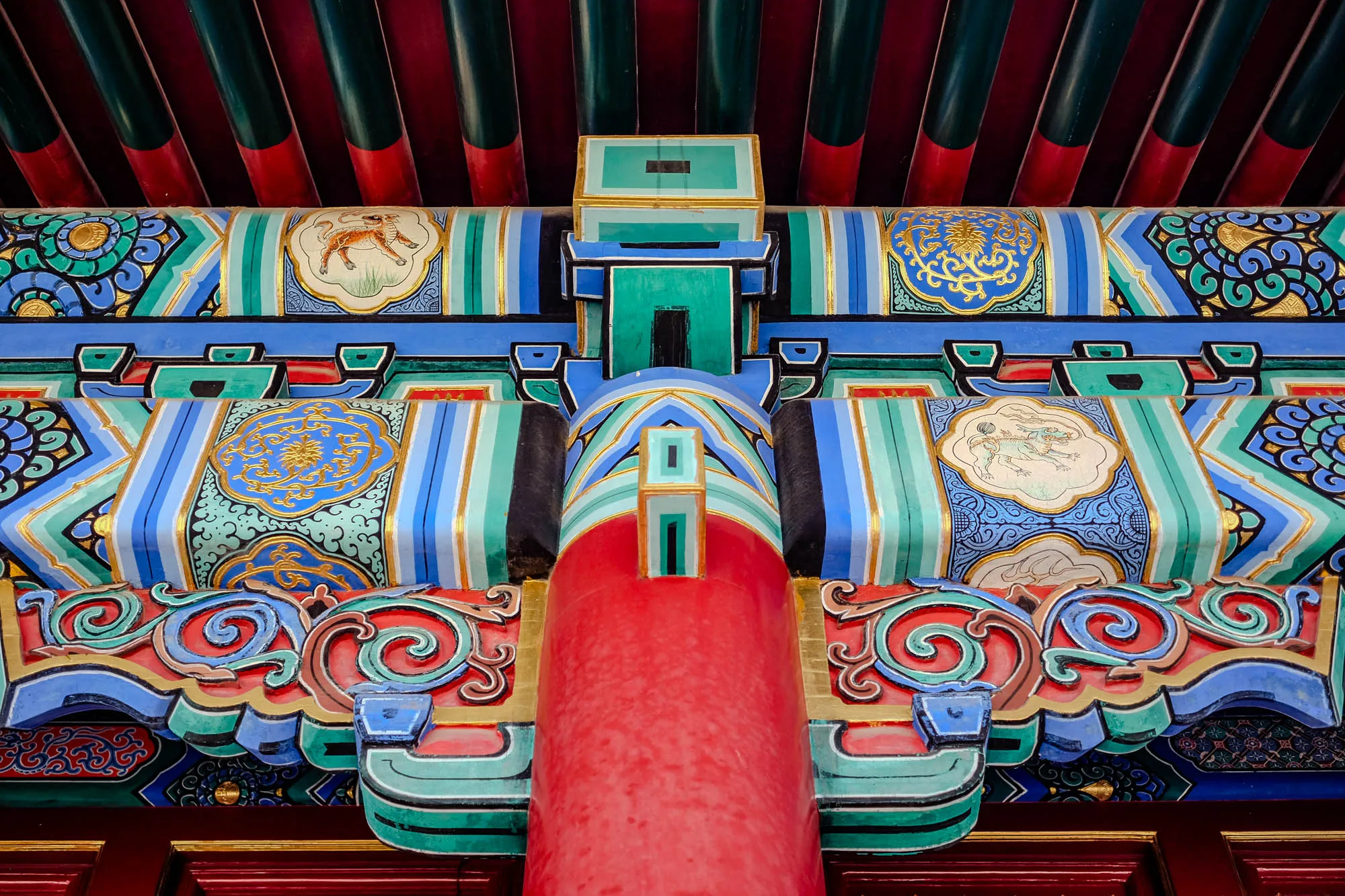 This is a close-up of a brightly colored, highly ornate ceiling detail of a Chinese temple. The ceiling is comprised of many rows of horizontal, parallel beams painted in alternating shades of red and green. Above this is a section of carved and painted wood, with an intricate design in shades of red, green, blue, gold, and white. This section is centered over a large, bright red pillar with a few more carved details. The carved wooden details are primarily a swirling, organic pattern with a few animal figures interspersed.  The image appears to be cropped, making it impossible to see the full extent of the ceiling or the pillar.