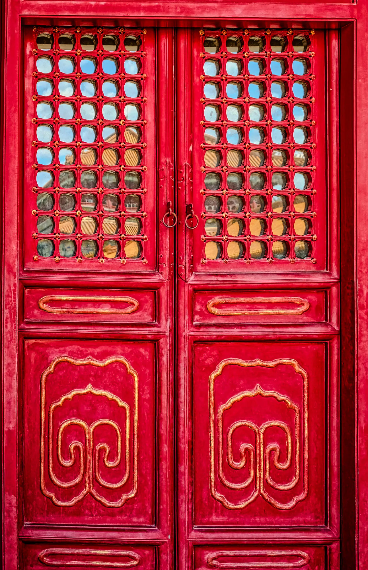 The image shows a pair of red wooden doors with intricate gold detailing. The doors have a lattice window on the top half. The lattice is made of a grid of small, round holes, with a gold trim around each hole. Behind the lattice, you can see a reflection of the sky and some other buildings. The bottom half of each door features a large, gold, swirling design. There is a single, gold, ring-shaped handle on the right door. The background is also red, suggesting the door is part of a larger building.