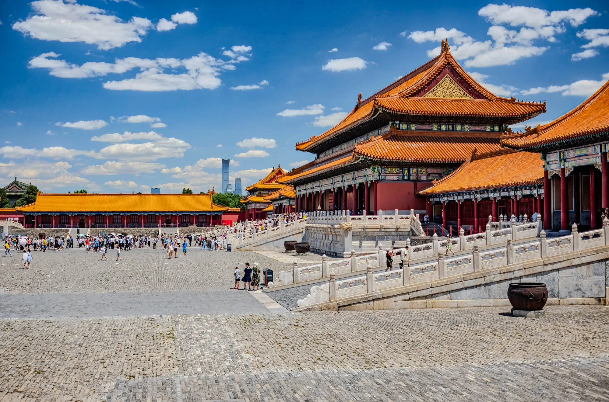 The image shows a view of the Forbidden City in Beijing, China. In the foreground, there is a large, paved courtyard with a few people walking around. On the right side of the image, there are white stone steps leading up to a large, red and gold building with a tiled roof. The building is decorated with intricate carvings and paint. There are also a few people walking on the steps and along the building. Behind the building, there is a blue sky with white clouds. In the distance, you can see a few more buildings with tiled roofs and the tops of some tall buildings.