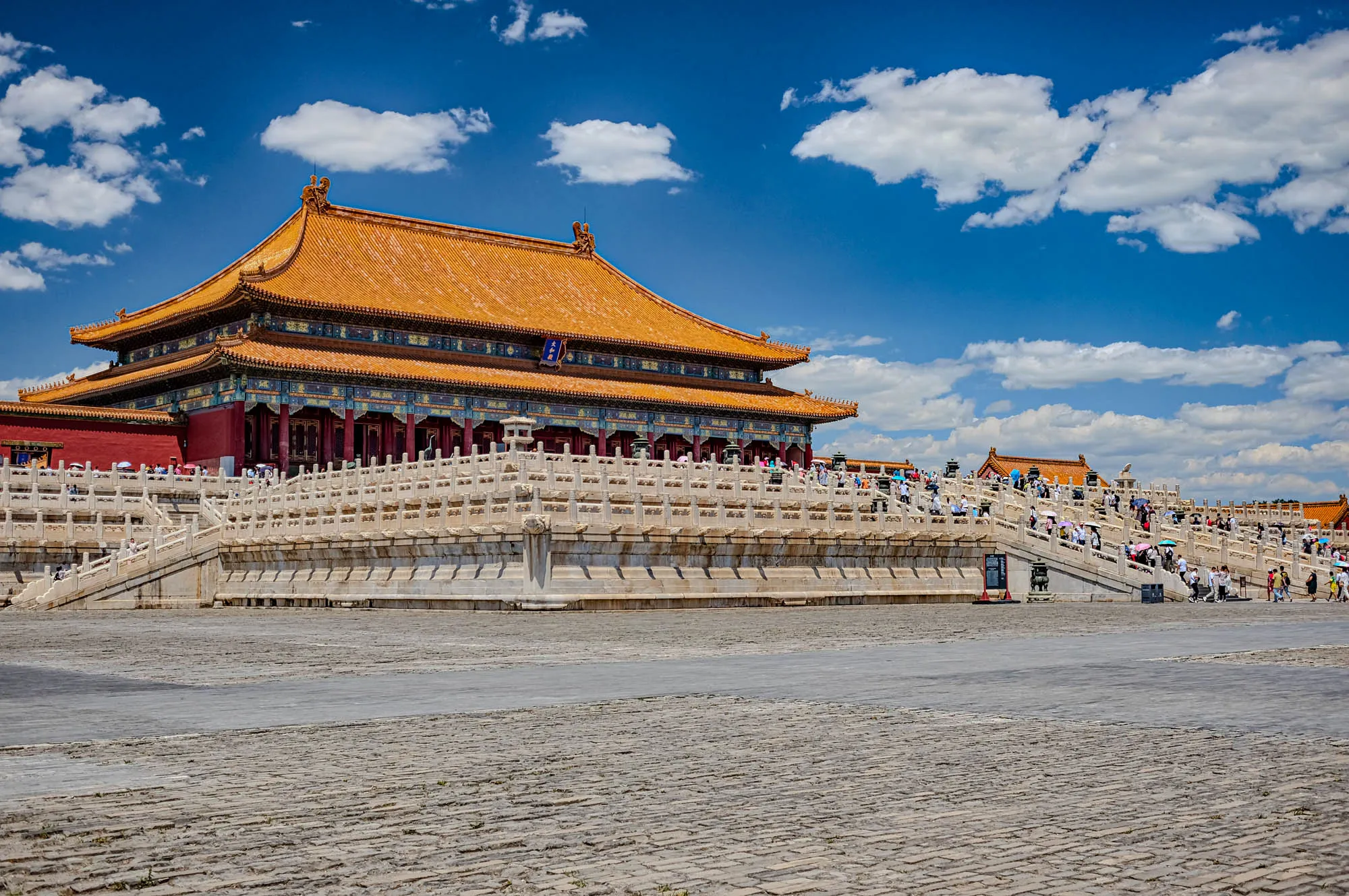 This is a wide-angle shot of the Forbidden City in Beijing, China. It shows a large palace building with golden tiled roof and red walls, set back on a raised platform with white stone balustrades and stairs. The palace is ornate with many decorative elements and has multiple levels. There are a lot of people walking around on the grounds. The foreground is a large, open plaza with gray cobblestone paving. The sky is a bright blue with a few puffy white clouds.