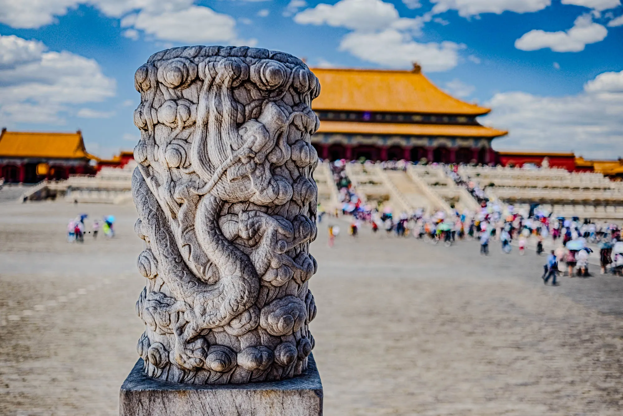 The image shows a close-up of a stone pillar with a dragon carving on it. The pillar is made of a gray stone and the dragon is carved in intricate detail. The dragon's head is not visible in the image. It is a side view of the pillar, and the dragon appears to be wrapping around the pillar, its body is made up of what appear to be many layers of scales and cloud-like details. The pillar is resting on a square base, and the background is out of focus, but a large building with a yellow tiled roof can be seen in the background. There are many people blurred in the distance walking in front of the building. The sky is blue with white fluffy clouds.