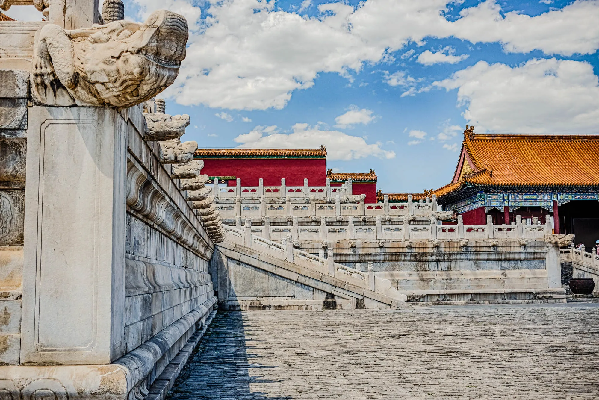 The image shows a stone wall and staircase leading up to a red and gold building with a tiled roof. The wall is carved with intricate designs and has several gargoyle-like sculptures, one of which is in focus in the left foreground. The building in the distance has a red wall and gold trim, with a tiled roof in a more traditional Chinese style. The image is taken from a low angle and the background is a bright blue sky with white clouds. The stone steps and pavement are gray and worn, suggesting age and use. The overall impression is one of grandeur and antiquity, with a hint of mystery and intrigue. The scene is likely a part of the Forbidden City in Beijing, China.