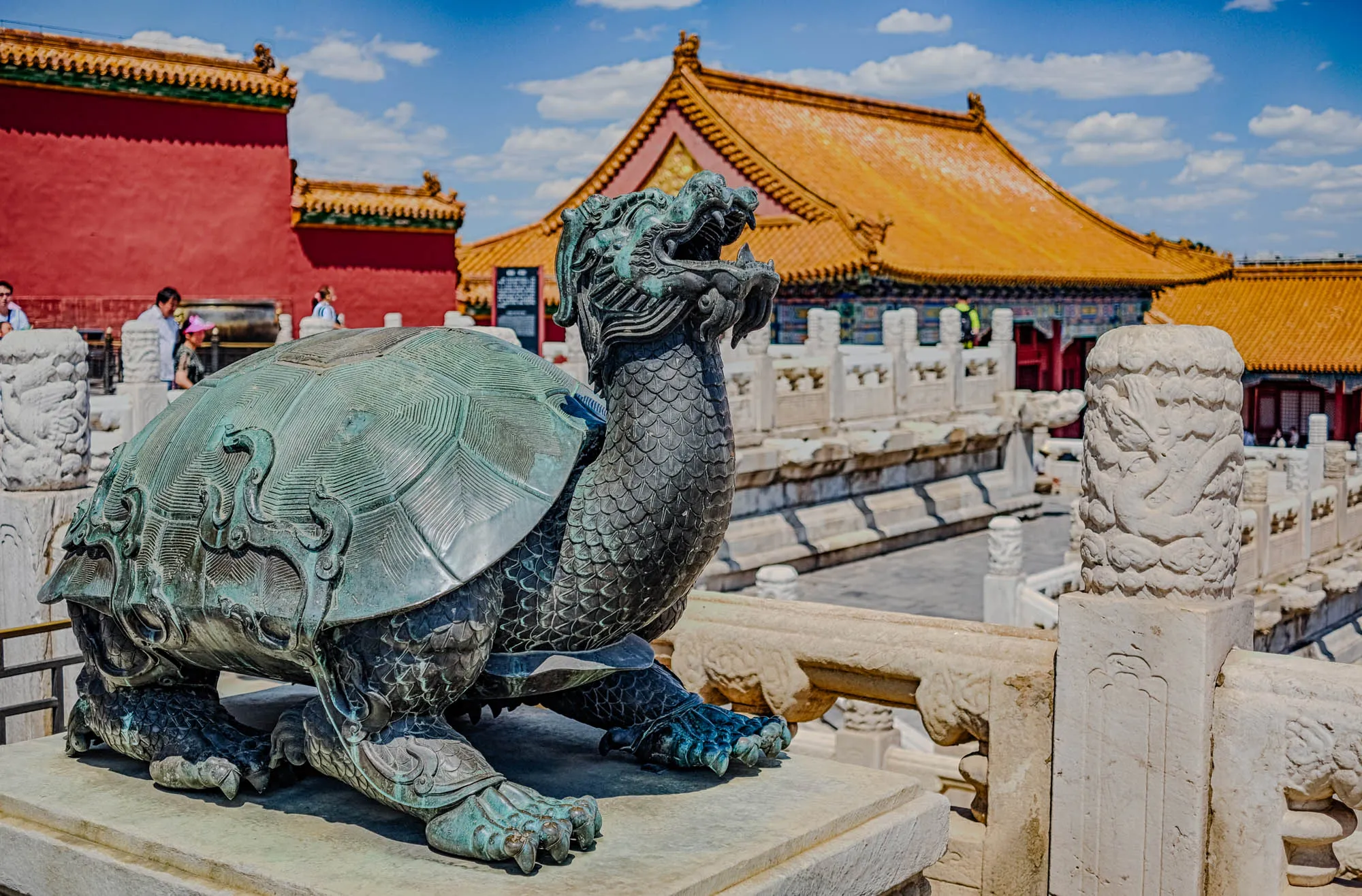 The image shows a bronze statue of a turtle with a dragon head in front of a large building with a red and gold roof. The turtle is facing the right side of the image and is lying on a stone platform. The turtle is detailed with intricate carvings, including scales on its shell and claws on its feet. The dragon head is also detailed with scales and sharp teeth. The building behind the turtle is decorated with a red and gold roof and has a white stone fence in front of it. The background is a blue sky with some white clouds. There are a few people visible in the background near the building. The overall feeling of the image is one of peace and tranquility. The bronze statue of the turtle and dragon is a symbol of good luck and fortune in Chinese culture.