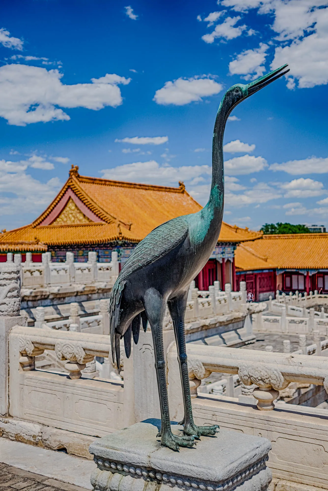 The image shows a bronze statue of a crane standing on a stone pedestal in front of a white marble railing. The crane has a long, thin neck that is bent back at an angle. The statue is weathered and has a greenish tint. Behind the statue, there is a building with an orange roof and white walls. The background of the image is a blue sky with white clouds.