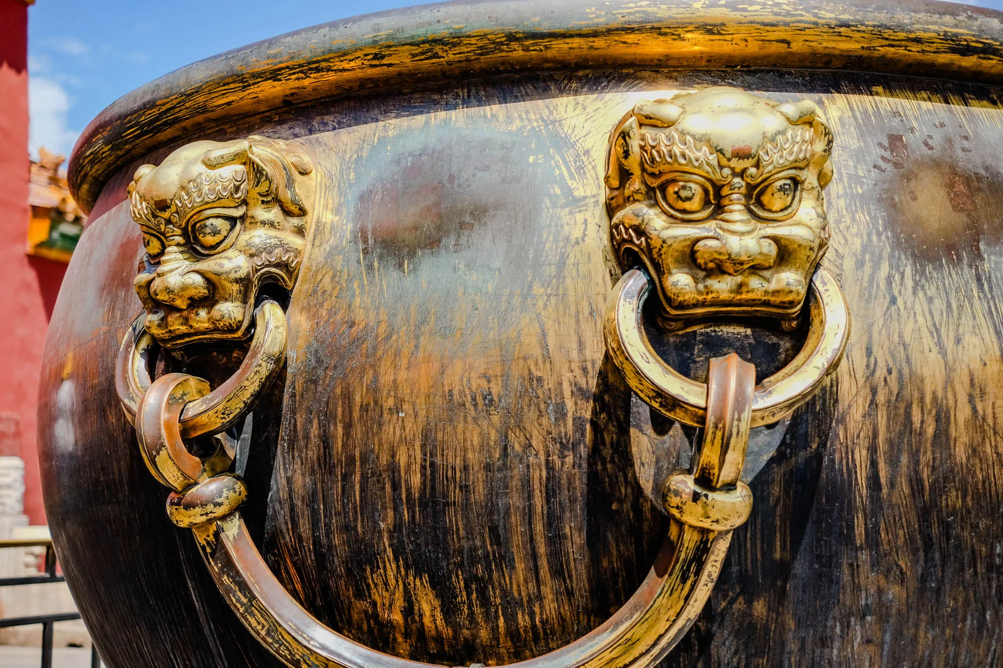 The image shows a close up of a large bronze vessel, likely a ceremonial gong or drum. It has two ornate handles, both shaped like the face of a snarling mythical beast. The handles are made of gold, with intricate details carved into the fur and face. The beast's mouth is wide open, showing sharp teeth. The vessel itself is made of dark bronze, with a patina of age and use. It has a smooth, rounded surface, with fine lines of wear and tear etched into the metal. The image is likely taken outdoors, as there is a blue sky visible in the background. The vessel is large and imposing, suggesting it was used for important rituals or ceremonies. It's a powerful image, showing both the beauty and the power of ancient Chinese art and culture.