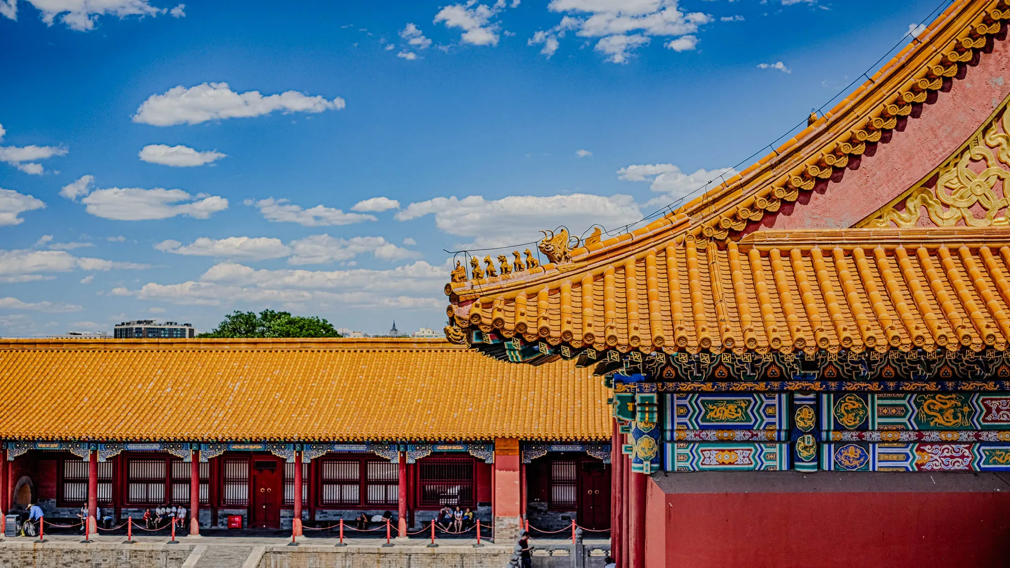 The image shows the corner of a Chinese temple or palace, with a bright blue sky and fluffy white clouds above. The corner of the roof is decorated with golden dragons and other ornate details. The roof is a bright yellow, and the temple walls are red. The lower portion of the image shows a section of the temple with more yellow roof, red columns, and a grey tiled floor. There are a few people walking around the temple grounds, but the image is mostly focused on the architectural details of the building.