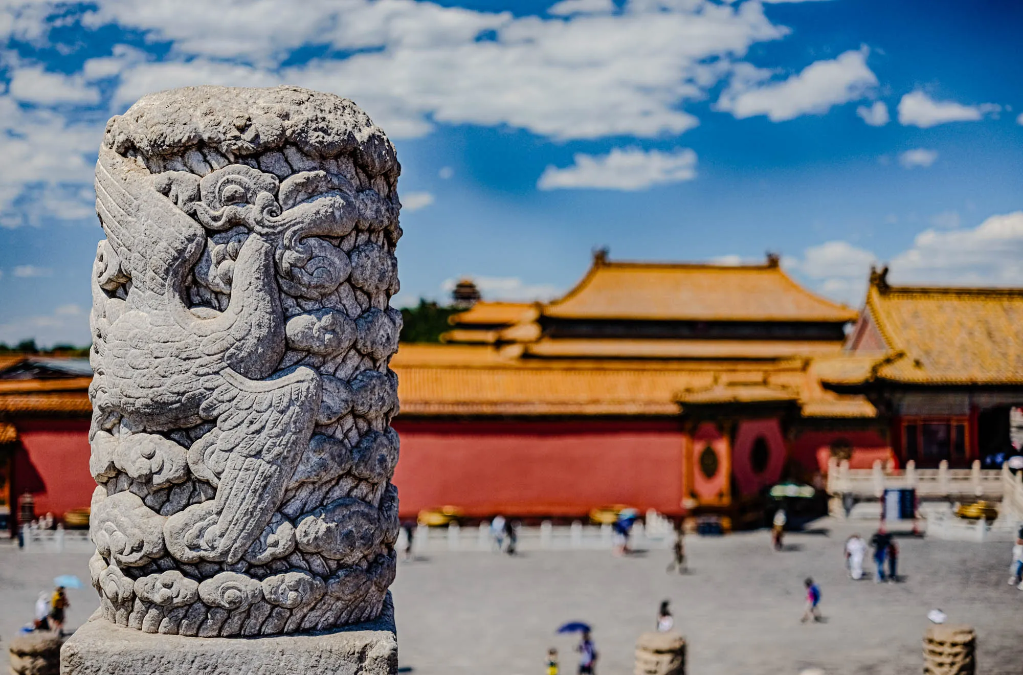 The image is a close-up of a stone pillar, carved with the image of a mythical bird. The bird's wings are spread out, and its head is turned to the side. The pillar is in front of a large, red building with golden roof tiles. The building is out of focus, suggesting that the pillar is in the foreground. The sky is blue and there are white clouds in the background. The scene evokes a sense of history and grandeur. There are people walking around the courtyard, a few holding umbrellas to shield themselves from the sun.  