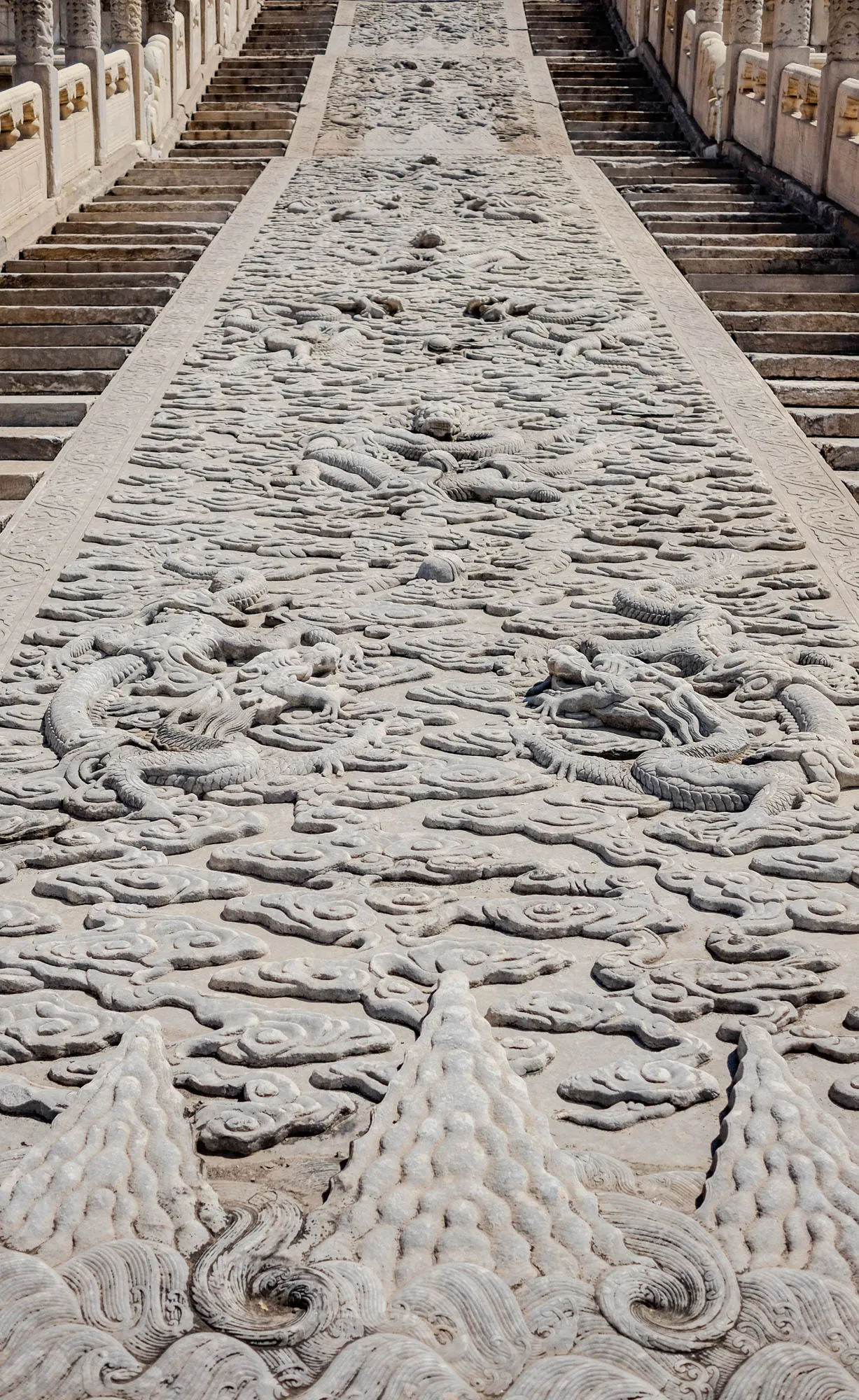 The image shows a long, straight pathway leading up to a set of stairs. The pathway is made of stone and is covered in intricate carvings. The carvings are of dragons and clouds. The pathway is flanked by two sets of stairs, each with a stone railing. The stairs are made of rough-hewn stone and are worn from years of use. The stonework is weathered and gray. The perspective is looking down the pathway, with the stairs receding into the distance. There are several carvings of dragons on the pathway, with their bodies and scales clearly visible. The clouds are carved in a flowing, wispy style. The entire scene is a testament to the skill and artistry of the ancient Chinese stonemasons.