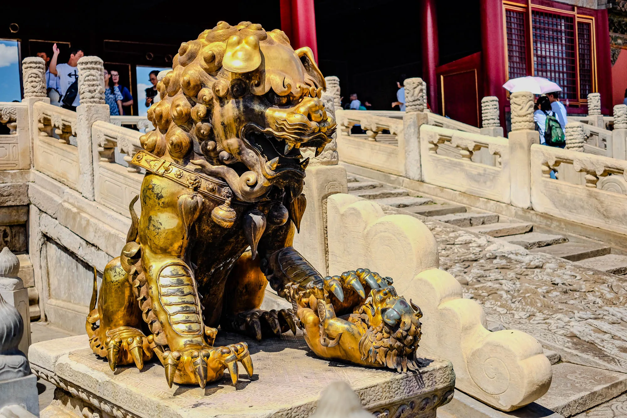 The image shows a large golden statue of a lion, or possibly a mythical beast like a griffin, sitting on a white stone platform. The statue is detailed, with intricate carvings and a curled mane. It's posed with one paw raised, holding a smaller golden sculpture that appears to be a smaller lion. Behind the statue, the platform extends back into a series of steps and an elaborate stone railing, suggesting a temple or palace setting. In the background, people can be seen moving about, but they're obscured by the architecture and the statue. The overall impression is one of grandeur and ancient power, with the gold statue standing as a majestic guardian figure. 
