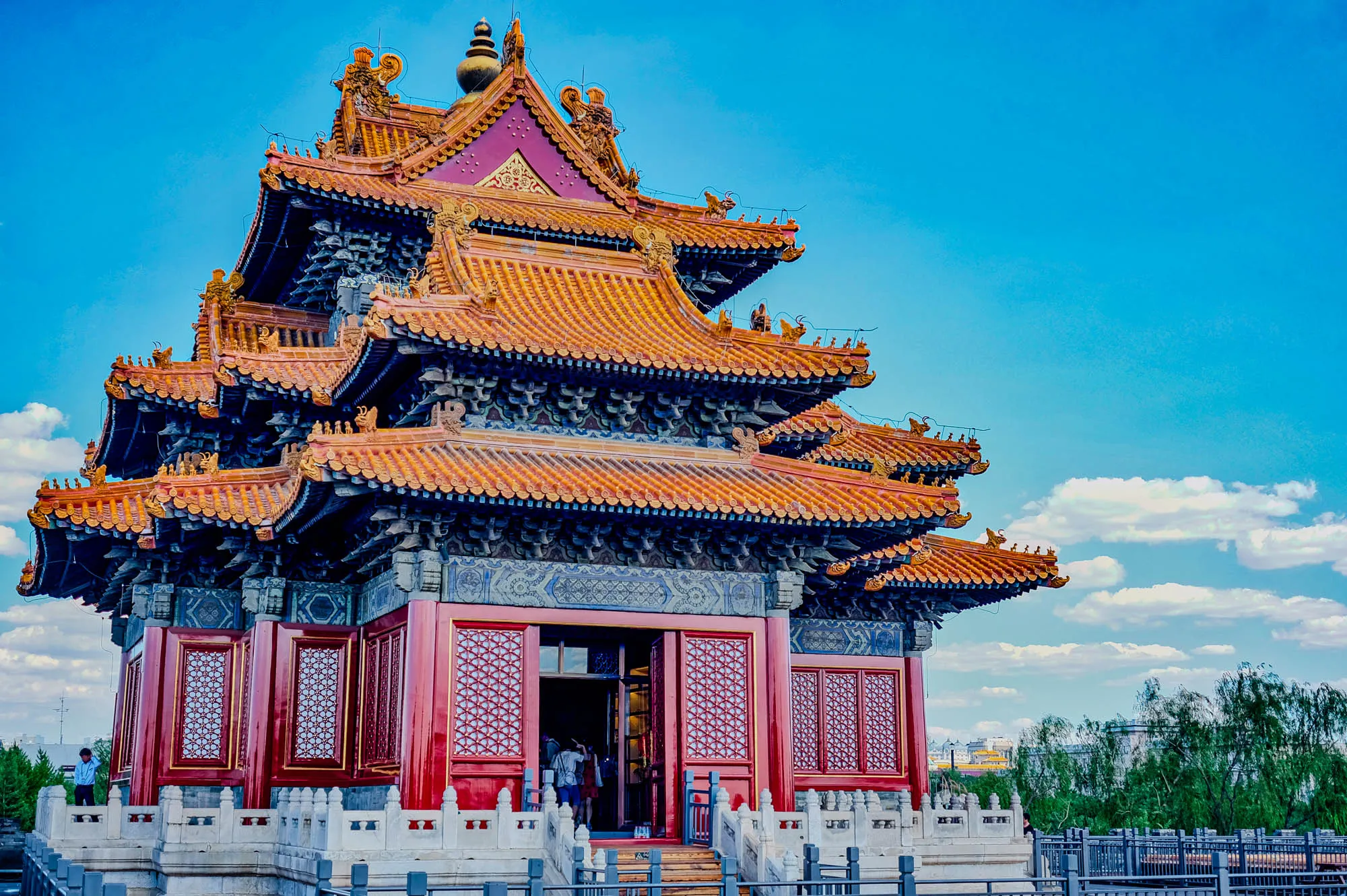 The image is a photo of a traditional Chinese building with an elaborate roof. The building has a multi-tiered roof with orange and black tiles and is decorated with intricate carvings and golden details. The building's walls are red and feature decorative windows with intricate patterns. There is a white stone railing around the building. There are people standing in front of the building, and some trees and a fence can be seen in the background. The sky is a bright blue with some white clouds.