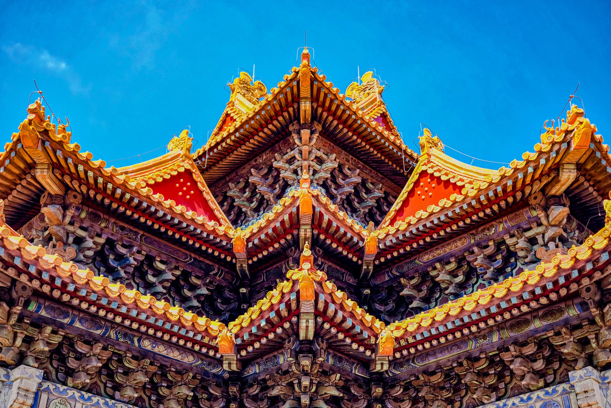 The image shows a close-up view of the intricate roofline of a traditional Chinese building. The roof is made of a series of layered eaves, each decorated with intricate carvings and paint. The eaves are painted in shades of red, yellow, and blue, and the carvings are made of wood. The roofline is symmetrical, with a central peak and two flanking peaks. The background is a clear blue sky. The roof is a beautiful example of Chinese architectural design.