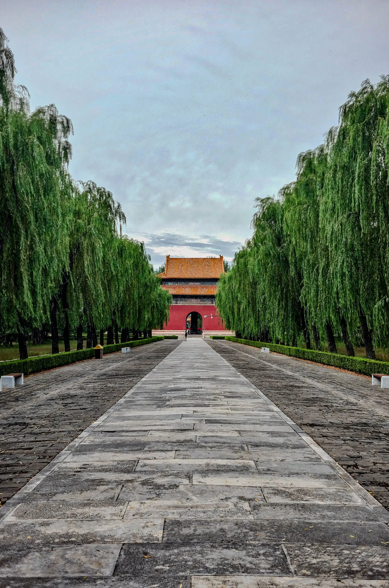 The image shows a long, straight path paved with grey stone. Two rows of tall, weeping willow trees line either side of the path. In the distance, there is a large red and orange gate with an arched entrance. The sky is a light grey with wispy clouds. The path is wide enough for two people to walk side by side, and there are two people visible walking towards the gate.