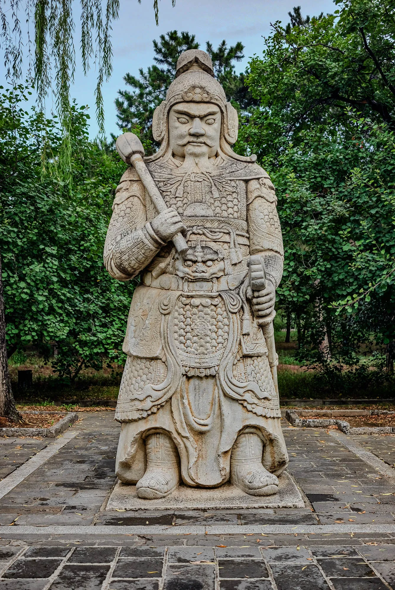 The image shows a stone statue of a warrior standing in a garden. The statue is carved in great detail and is wearing a helmet and armor. The warrior is holding a weapon in his right hand. The statue is standing on a stone base and is surrounded by green trees and bushes. The ground is made of brick paving stones. The statue is likely a representation of a Chinese warrior or general.