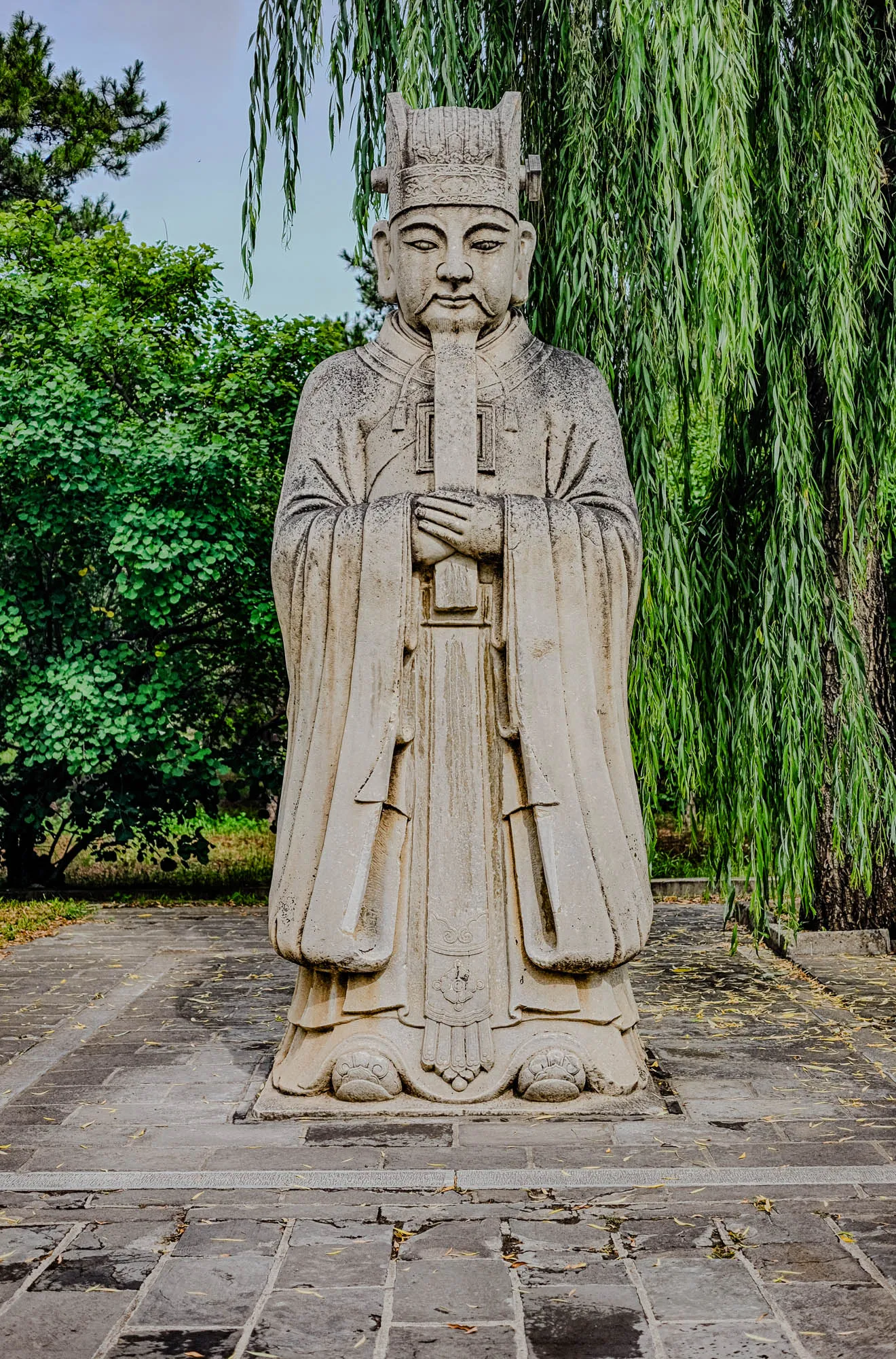 The image shows a stone statue of a man standing on a stone path in a garden. The statue is wearing a long, flowing robe and a tall hat. The man's hands are clasped in front of him. He has a stern expression on his face.  The statue appears to be weathered, with some moss and lichen growing on it. The background of the image is a lush garden with green trees and shrubs. There is a weeping willow tree to the right of the statue. The path leading up to the statue is made of large, flat stones.  The statue is set against a backdrop of lush greenery, which enhances its sense of age and tranquility.  It appears to be a traditional Chinese statue.