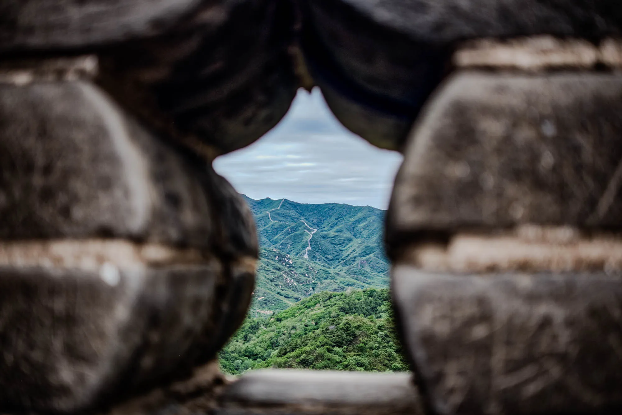 The image shows a view through a hole in a stone wall. The hole is shaped like a pointed arch. Through the hole, you can see a green, hilly landscape with a winding path snaking up the hill. The path is a light color and is likely made of stone or concrete. The sky above is cloudy. The stone wall surrounding the hole is made up of large, irregular stones.  