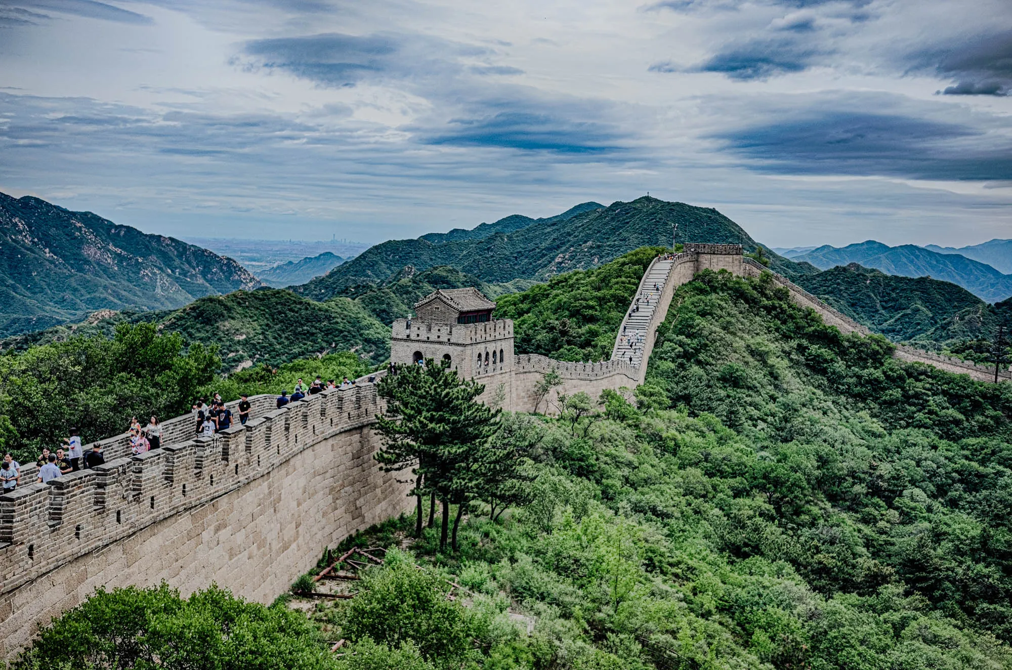 The image shows a section of the Great Wall of China, stretching across a mountain ridge. The wall is made of gray stone and is lined with a pathway. There are people walking along the pathway, some are going up, some are going down. The wall is surrounded by lush green trees and bushes. There is a small stone tower on the wall, with windows and a flat roof. In the distance, you can see a line of trees and buildings, which are likely part of a city. The sky is a light blue with some white clouds.