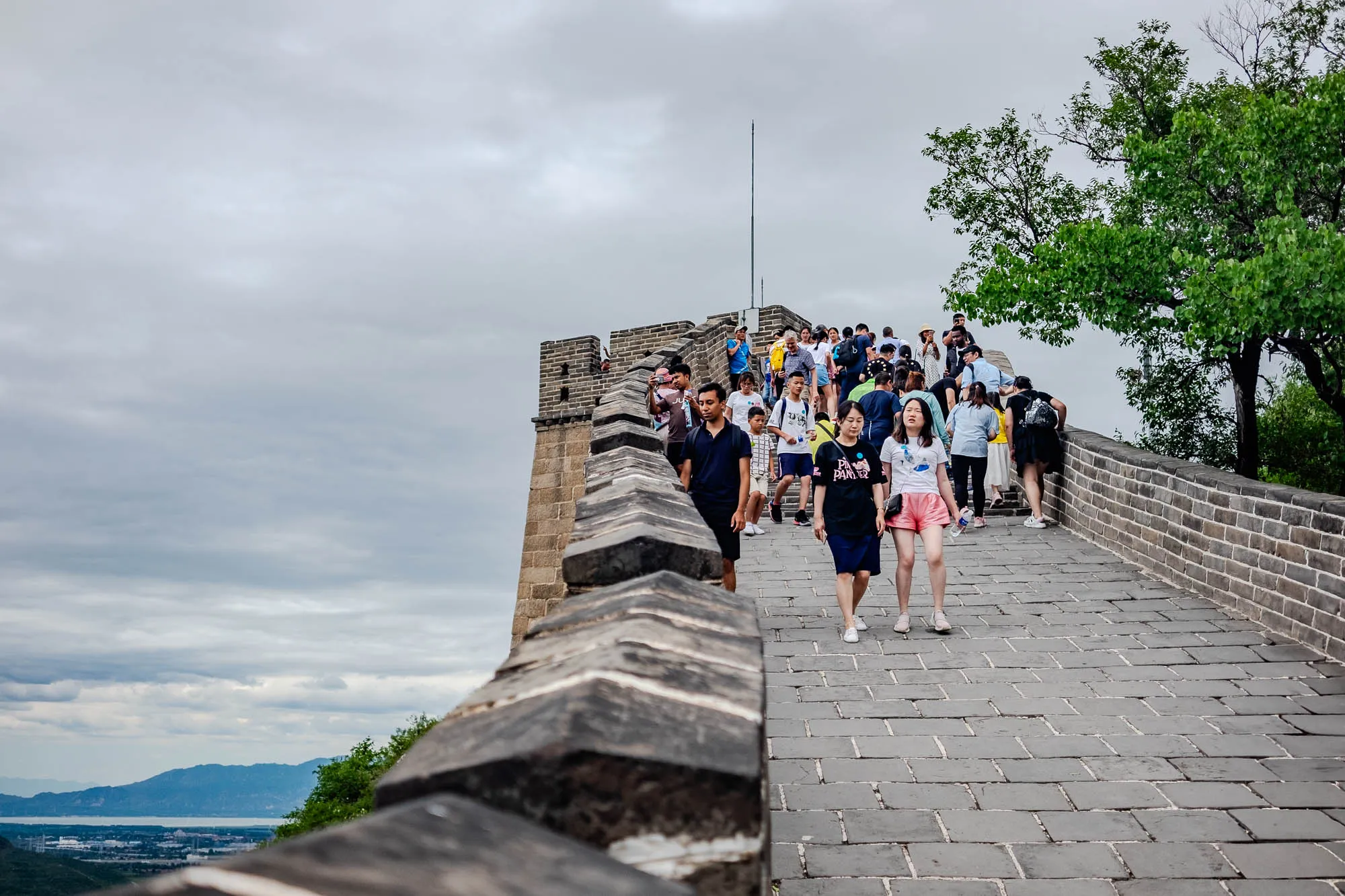 This image is a shot of the Great Wall of China. It is taken from a low angle, looking up the wall. The camera is focused on the paved path of the wall, where a group of people are walking. The people are mostly dressed in casual clothes, and some are taking pictures. The wall is made of gray stone, and there are trees growing on the hillside to the right of the path. In the distance, you can see a valley and a mountain range, as well as a small town or village. The sky is cloudy.