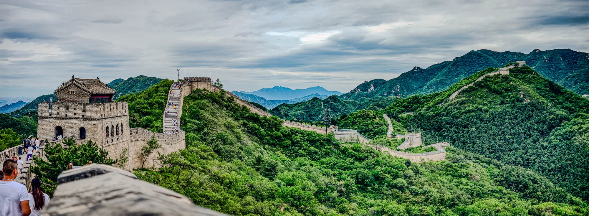 The image shows the Great Wall of China, with a large section of the wall in the foreground. It appears to be a watchtower with people walking on top of the wall. In the background, the wall snakes its way up a steep, forested hillside and disappears in the distance.  In the far distance, there are mountains on the horizon.  The sky is cloudy and grey. 
