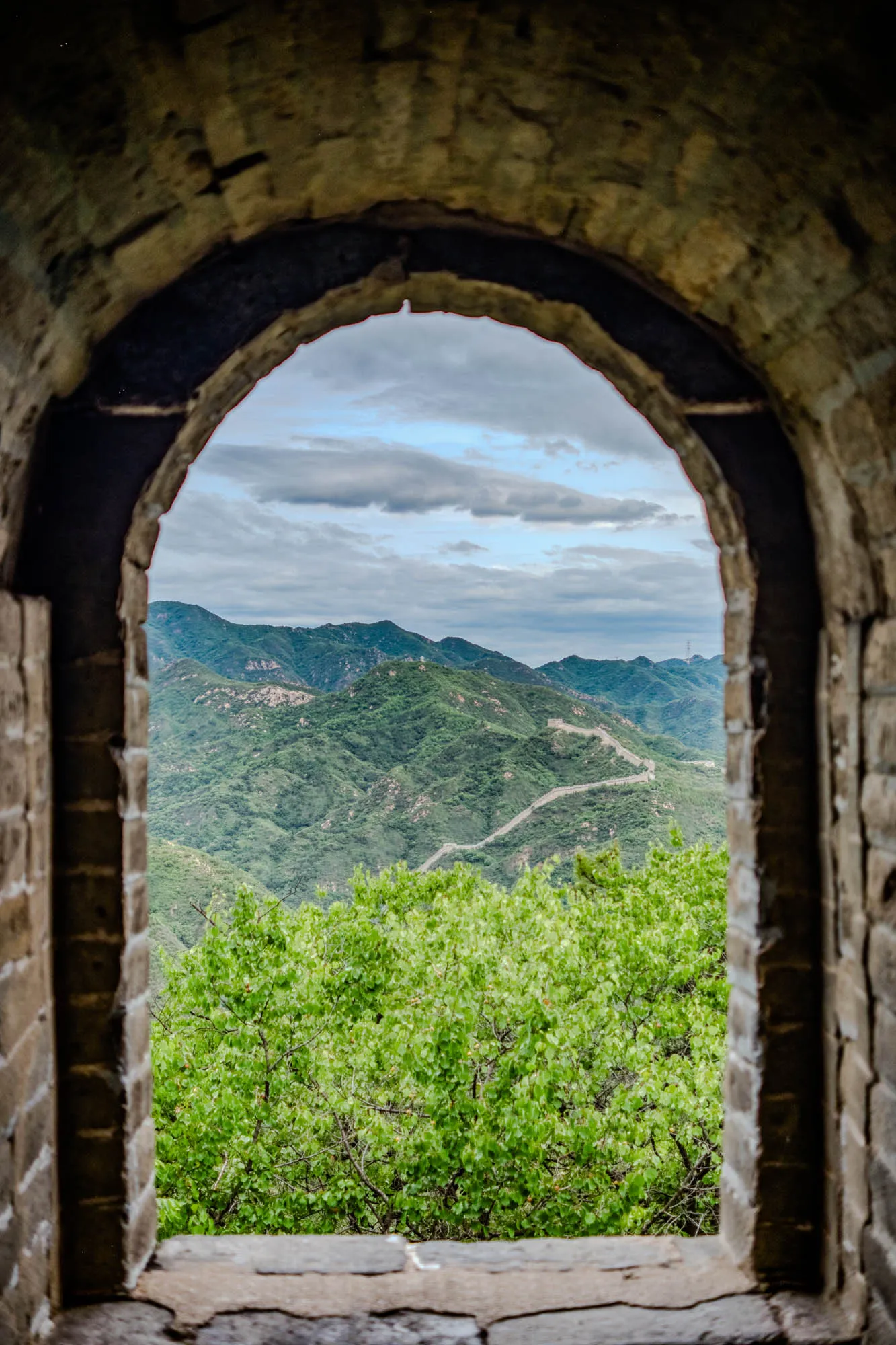 This image shows a stone archway that frames a view of rolling green hills and a blue cloudy sky. The archway is made of rough, weathered stone and appears to be part of a wall. The view through the archway shows a verdant landscape with green trees and shrubs in the foreground, followed by a series of hills that stretch back into the distance. The sky is a light blue, with a few puffy white clouds scattered across it. The image gives the impression of being a peaceful and serene place, with a sense of history and natural beauty.