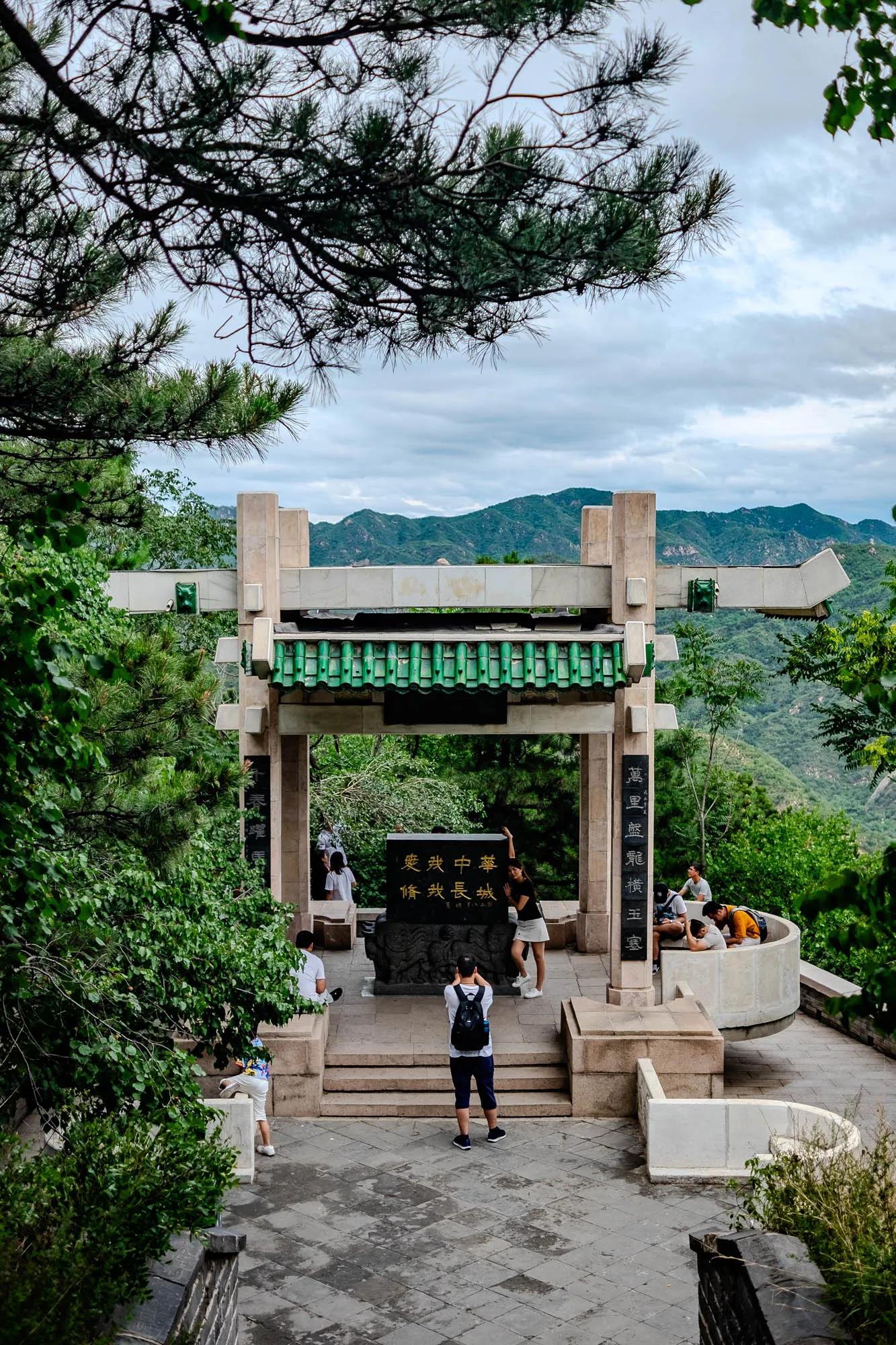 The image shows a traditional Chinese gate with a green tiled roof and white stone pillars. The gate is set against a backdrop of green hills. There are several people in the image. A man with a backpack stands near the gate, taking a photo. A woman in a white shirt and shorts stands by a black stone with Chinese writing. Several people are sitting on stone benches to the right of the gate. There is a large green tree in the foreground, obscuring the bottom of the image. The sky is mostly cloudy with a few areas of blue. The overall feel of the image is peaceful and serene.