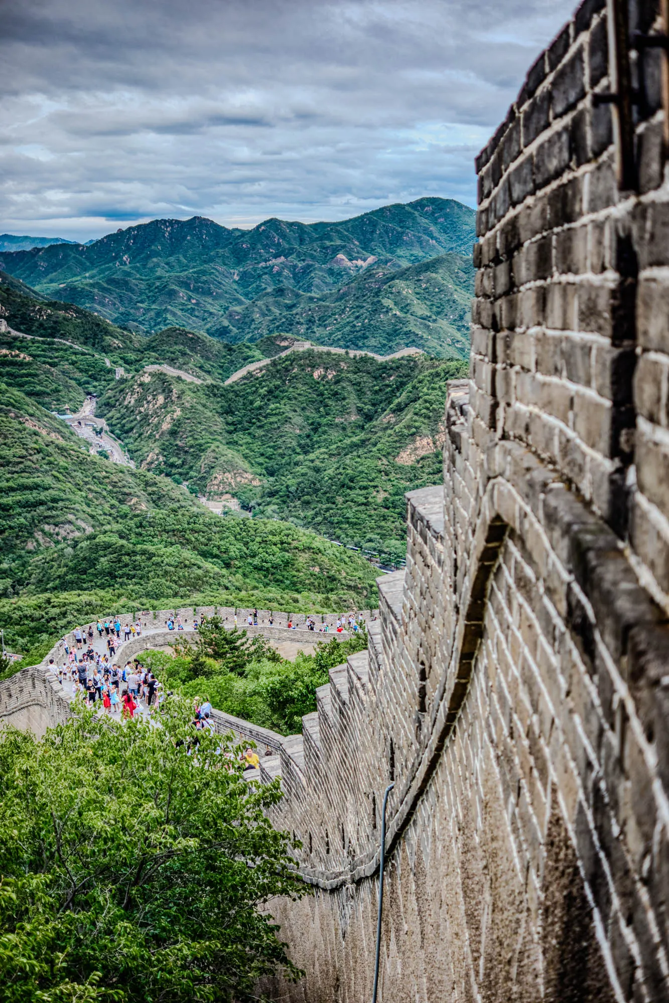 The image shows a close-up of the Great Wall of China. The wall is made of grey bricks and extends into the distance. The wall is built on a hillside with a long winding pathway. There are people walking on the wall, some are near the viewer and others are further away.  The image is taken from the perspective of someone standing on the wall looking down. In the background, there are rolling hills covered in lush greenery.  The sky is cloudy and the overall feeling is serene.  The image is a beautiful and iconic representation of this famous structure. 
