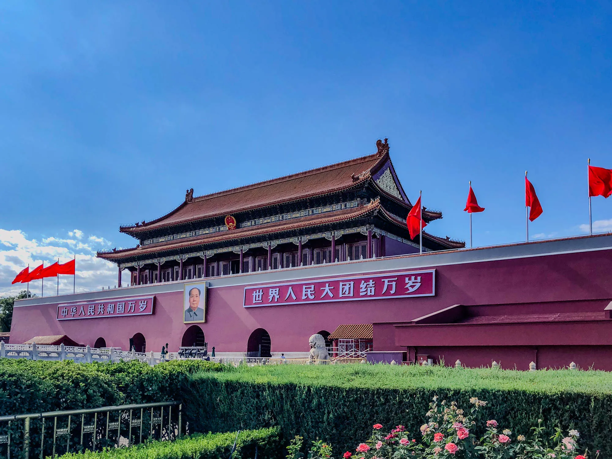 The image shows the Tiananmen Gate in Beijing, China. It is a large, red building with a traditional Chinese roof. The gate is flanked by two large, white stone lions. There are a few red flags flying on flagpoles outside the gate. There is a banner over the gate with Chinese writing, which reads: "Long live the people of the world." Below this is another banner in Chinese which reads: "The people of China stand united." There is also a portrait of Mao Zedong hanging on the wall of the gate. The building is surrounded by a wall, and there are green bushes and a small fence in front of the building. The sky above the gate is a light blue with a few clouds. The image was taken on a sunny day.