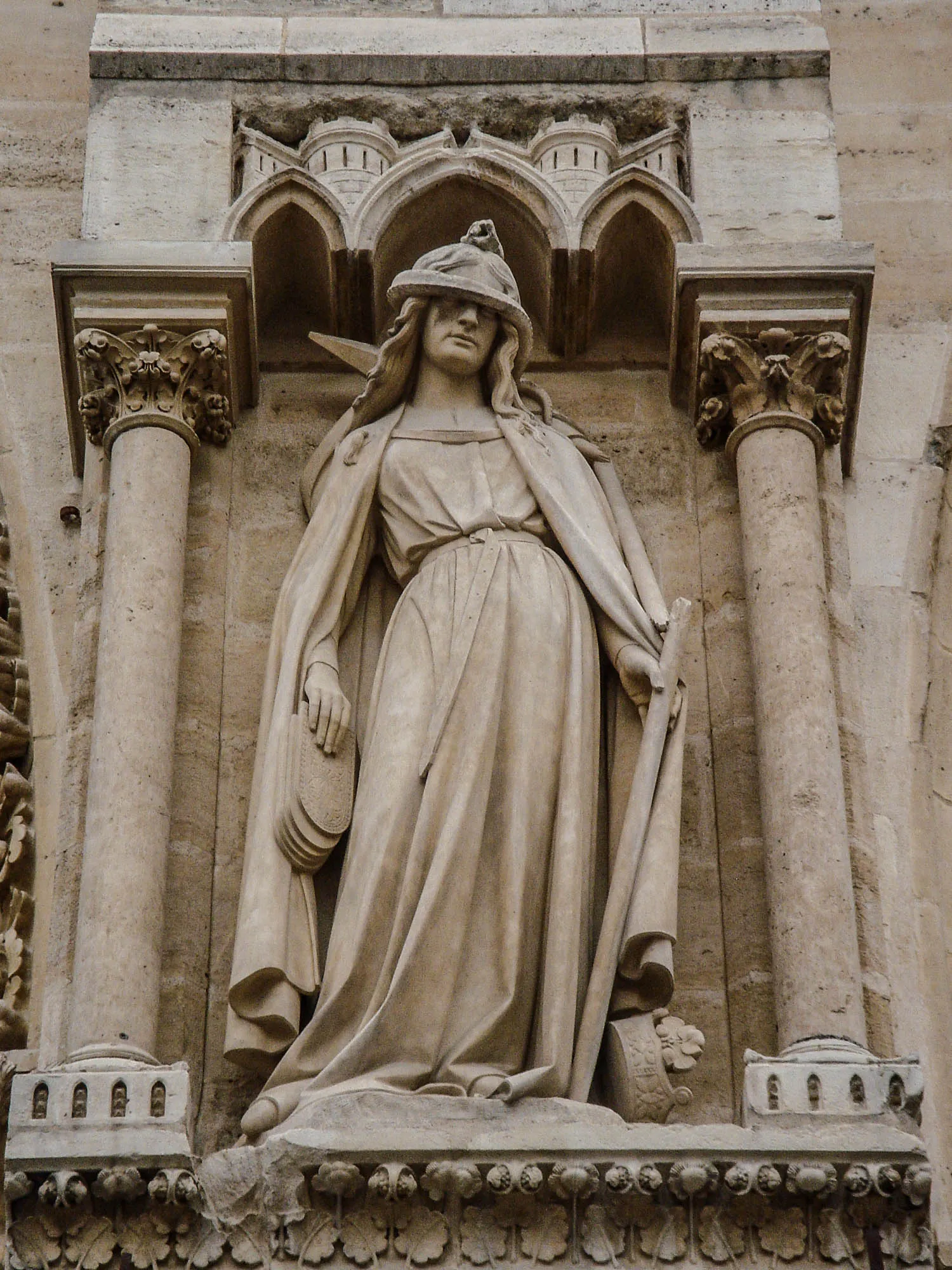 A statue of a robed women holding a broken staff in her left hand, tablets in her other, a crown at her feat, and a snake wrapped around her head covering her eyes.