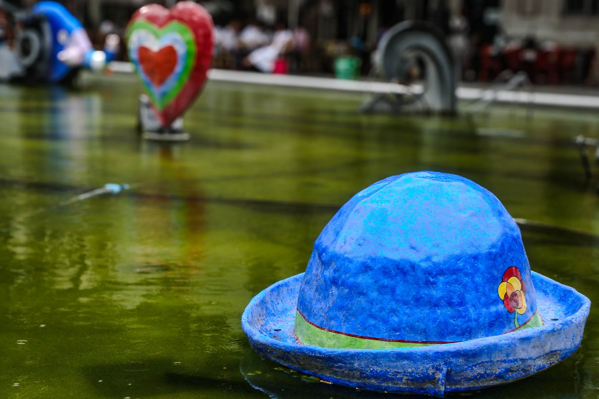 A sculpture of a bright blue bowler cap with a painted on gree band and abstract painged flower on the green water of a reflecting pool. A sculpted and painted heart, with bands of color going from red to green to blue, and back to red, is in the distance, along with two other scuptures that cannot be positively identified. All three are also on the water.