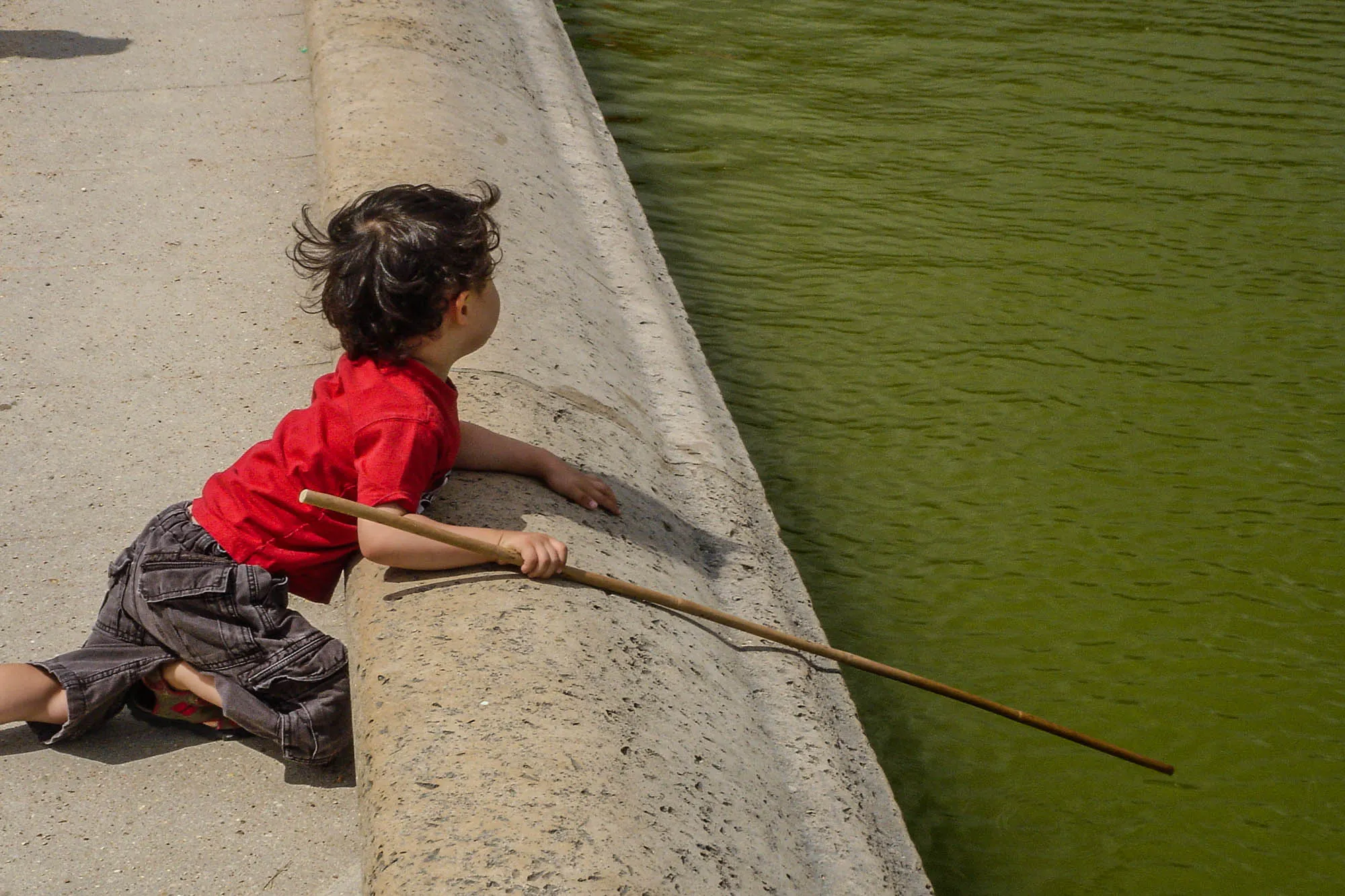 A young child leaning aginst the low retaining wall of a reflecting pool. They are holding a long stick and are wearing a red shirt and black washed out jean-like pants