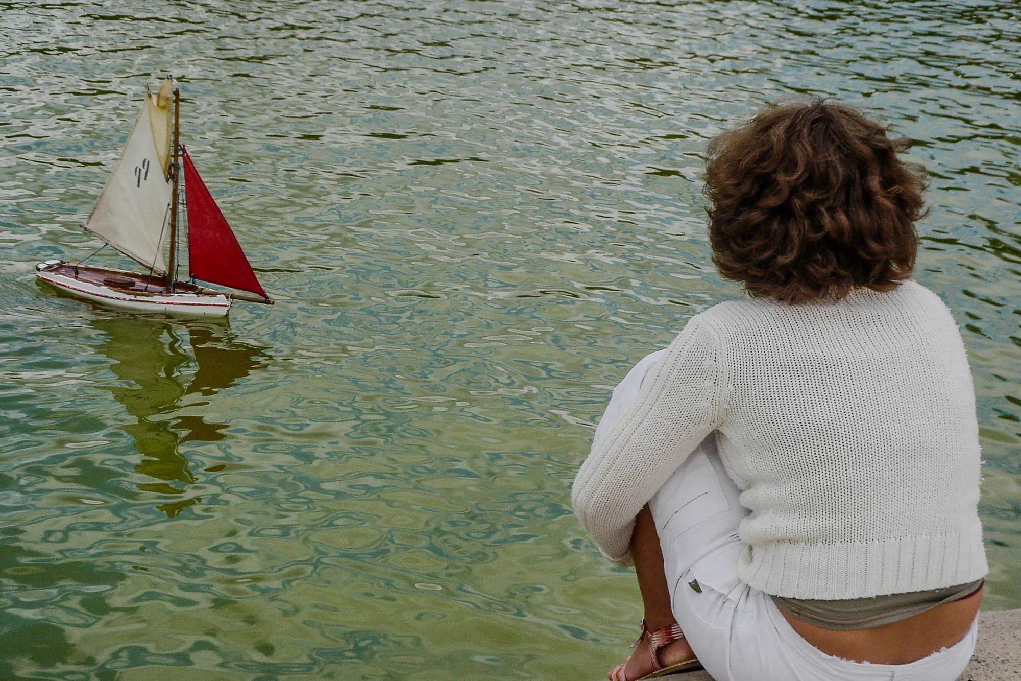 A young adult woman sitting on the edge of the retaining wall of a reflecting pool, her back to the camera, wearing a white sweater, white pants, and sandals. She's looking at a small sailboat in the pool.