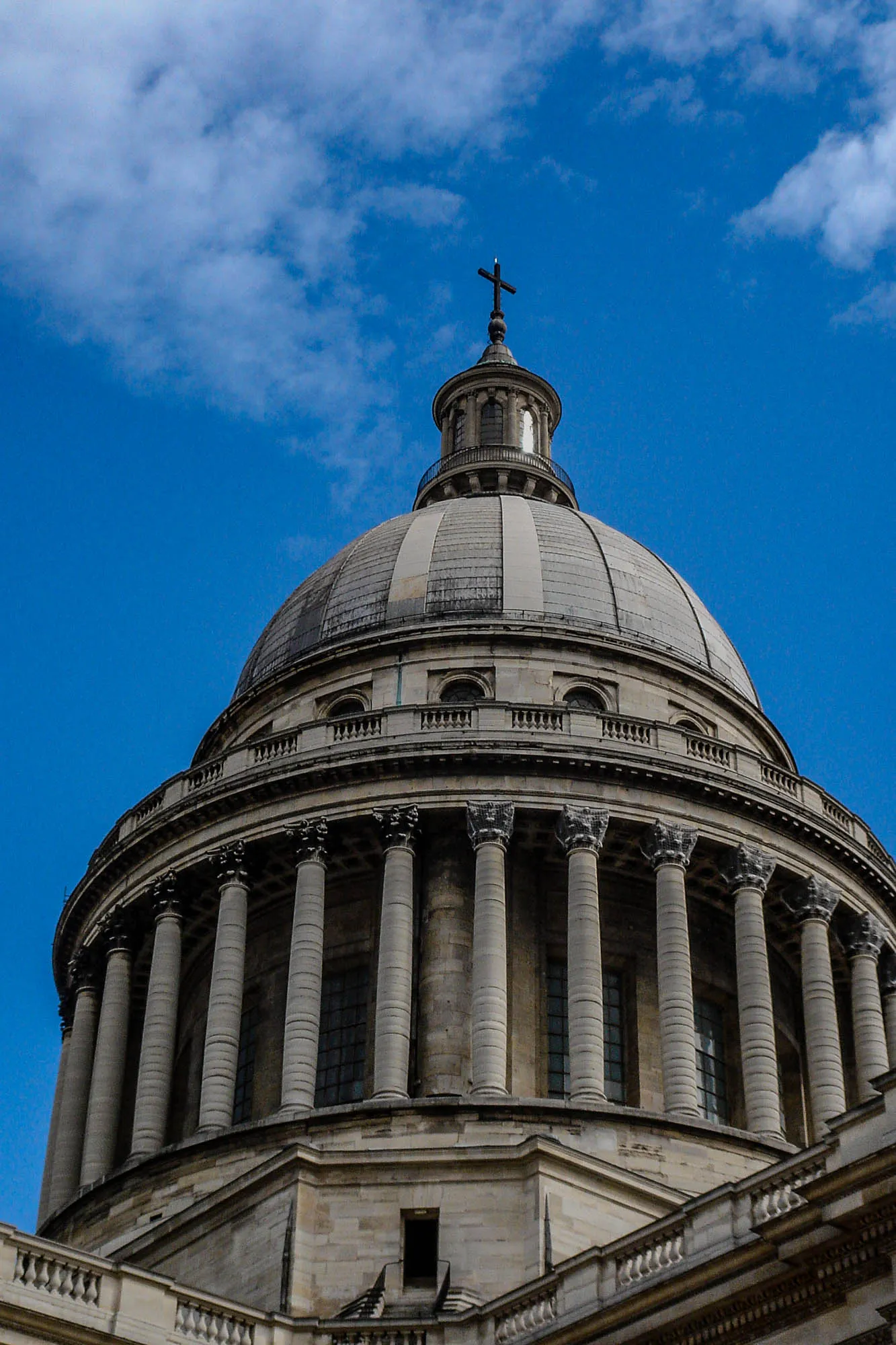 The stone exterior of a large round rotunda surrounded by pillars culminating in a dome with a roof lantern and a cross at the top.