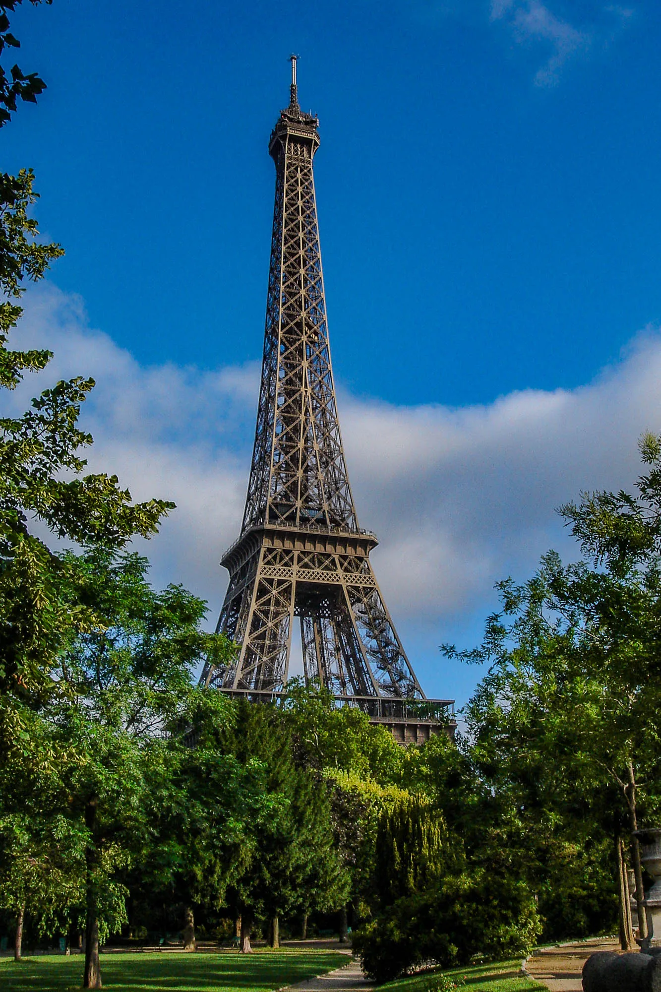 Eiffel Tower, emerging from behind trees in a park in front of it. The park has grassy areas and walking paths.