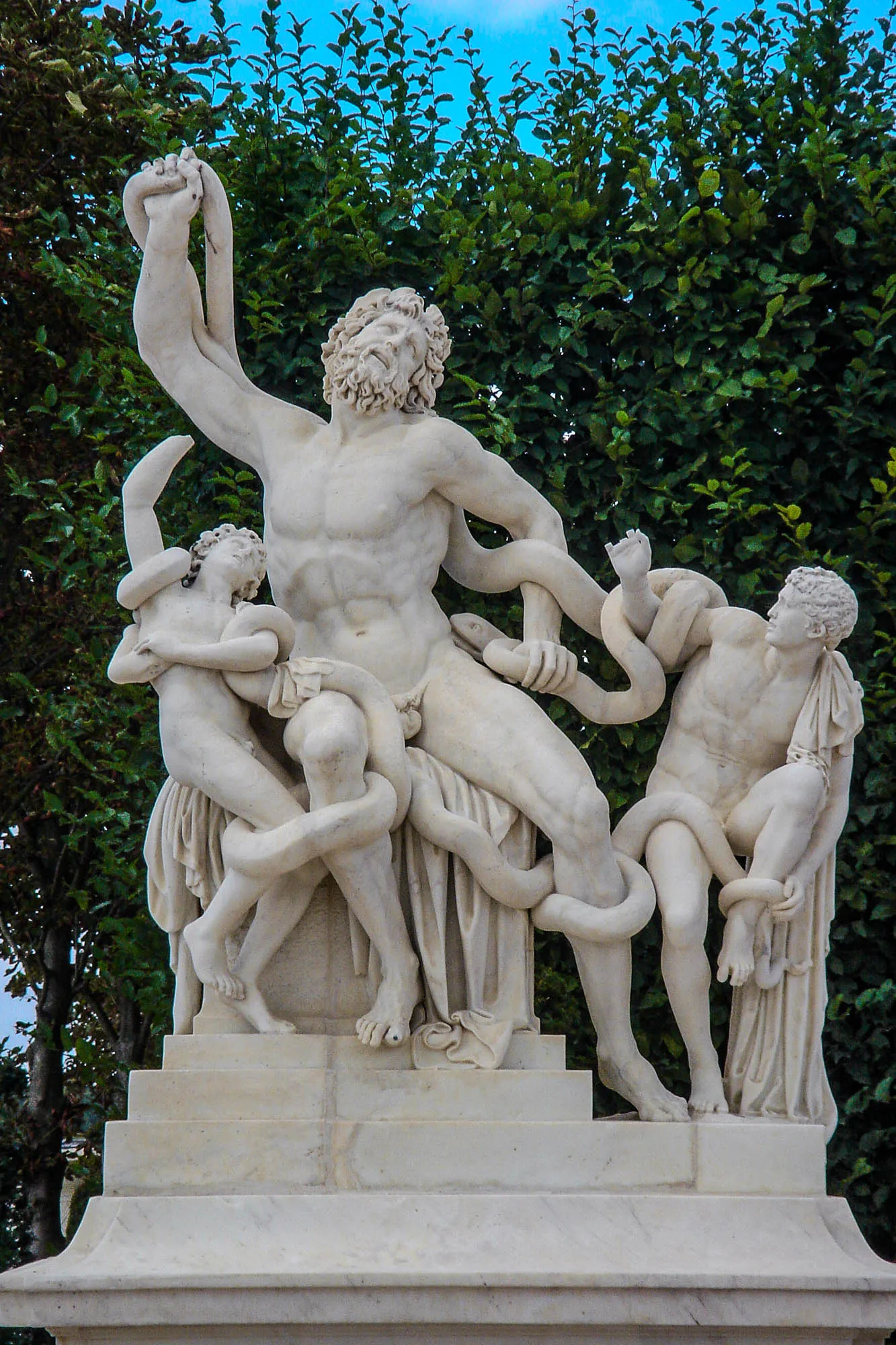 A marble statue depicting a man with a full beard sitting on a pedestal  and two young boys on either side. All three are struggling against a number of large snakes wrapping themselves around their legs, arms, and torsos.