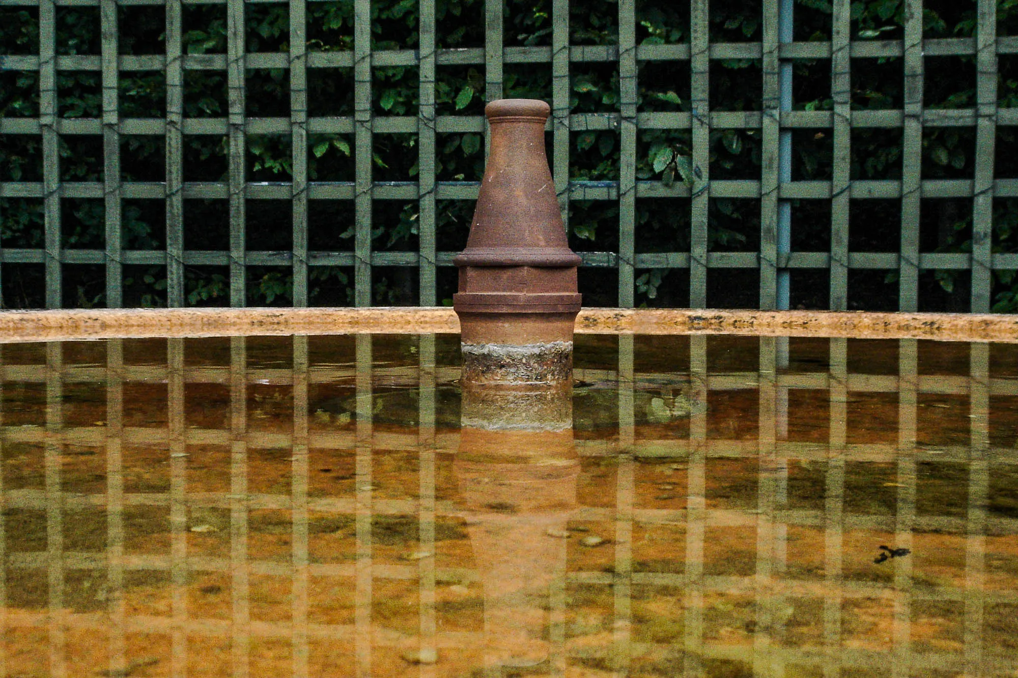 A green woodwn trellis reflects into the rusty water of a fountain's baisn. The fountain's nozzle, rusted and pockmarked, stands inoperable in the center.